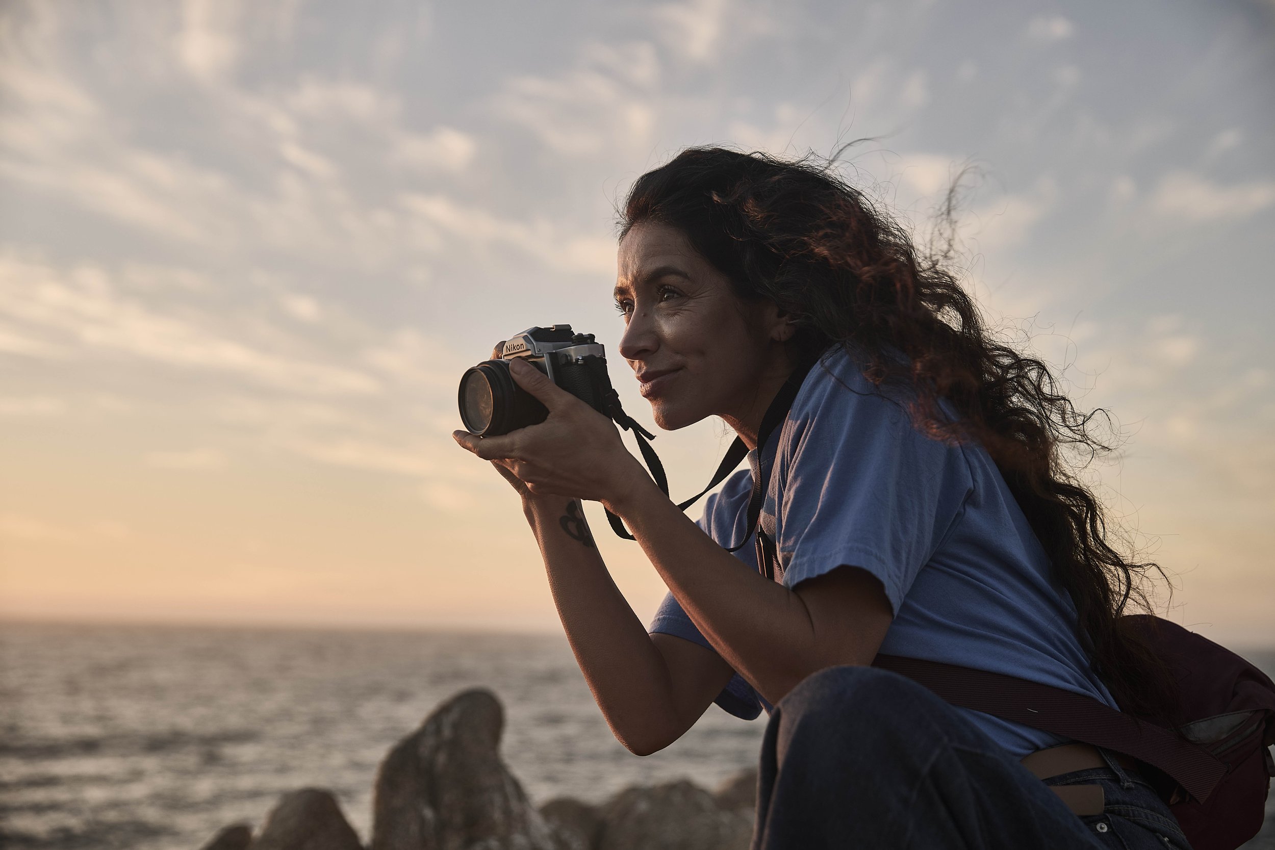 Mujer con cabello rizado, sosteniendo una cámara Nikon, en la orilla del mar durante el atardecer.