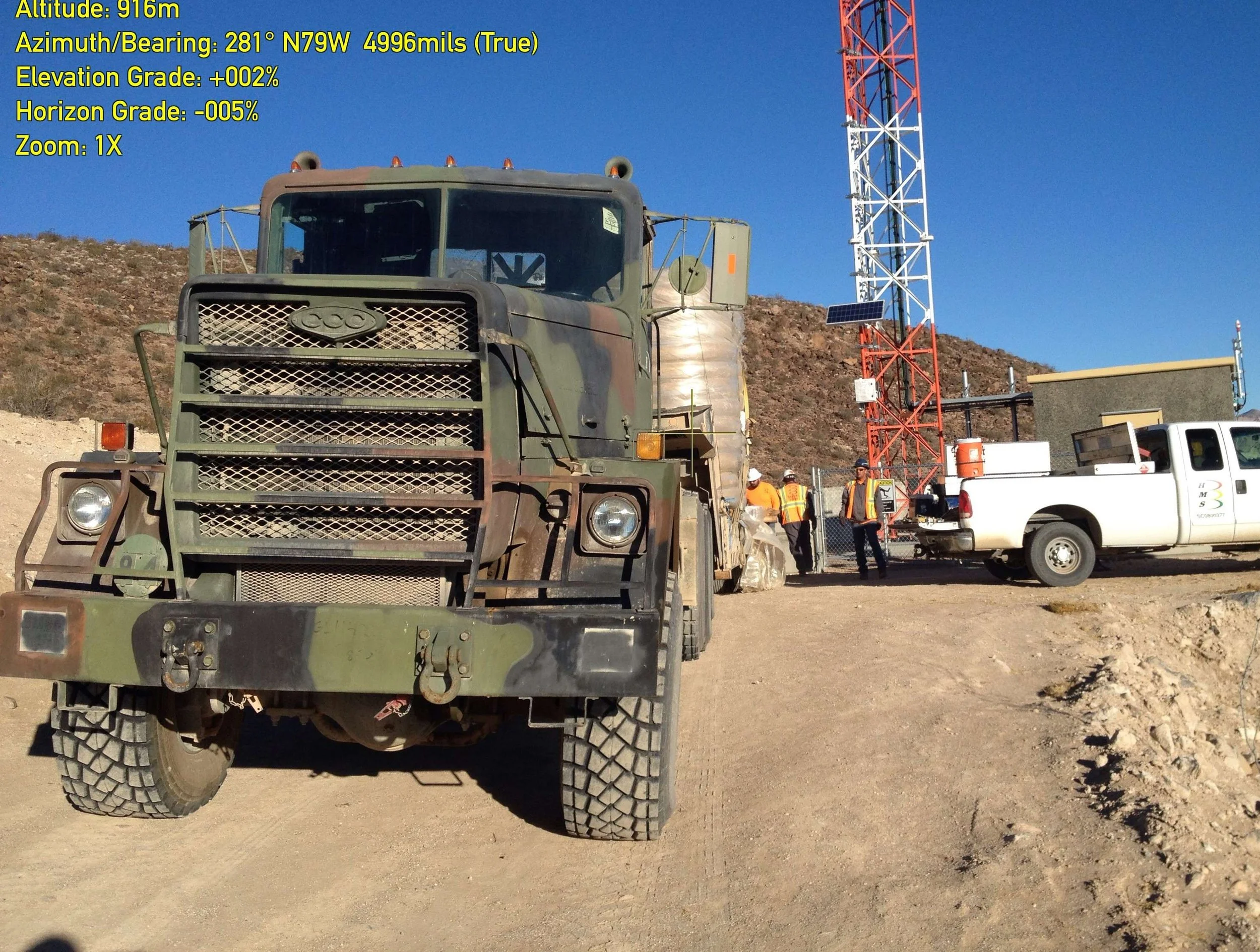 A green military truck parked on a dirt area with a large red and white radio tower and people in construction vests working nearby in the background. The sky is clear and blue.