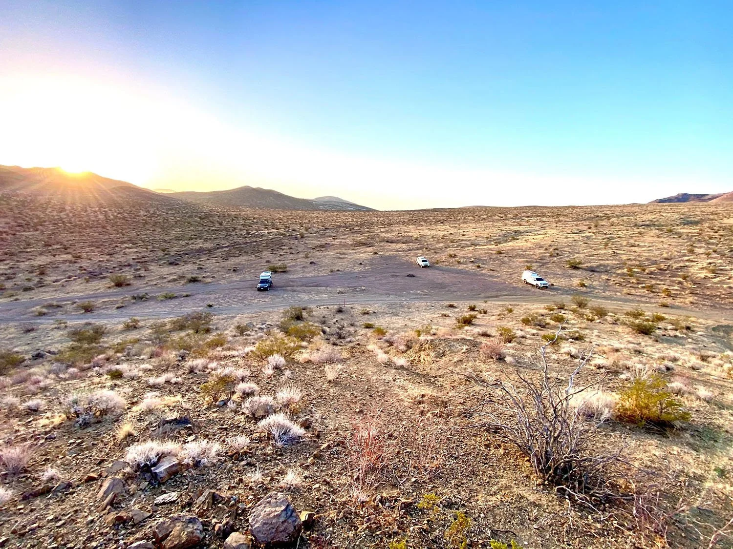 Desert landscape during sunset with three parked cars on a dirt parking area, sparse desert vegetation, and mountains in the distance.