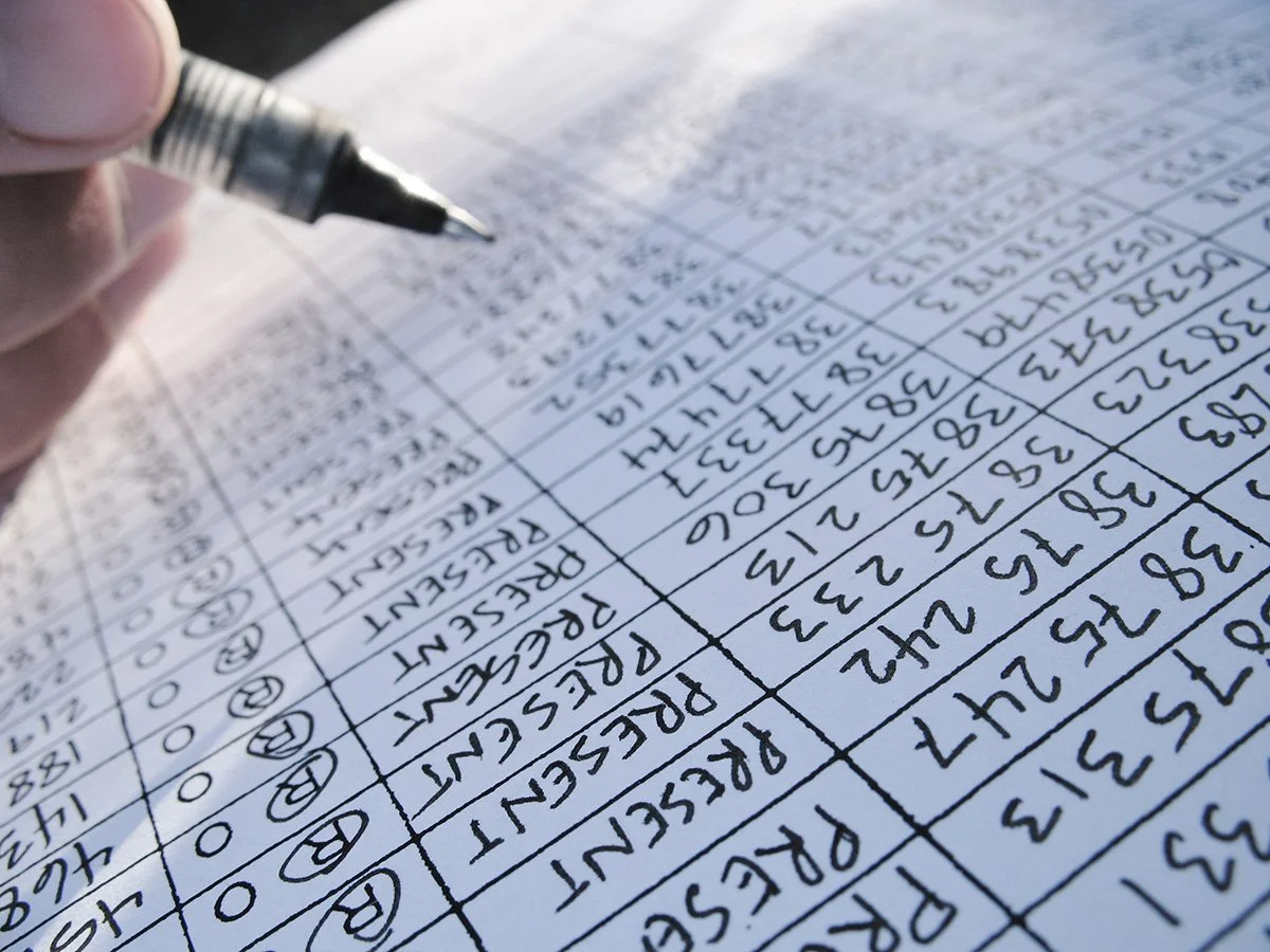 Close-up of a hand writing on a printed spreadsheet with a black pen, showing handwritten entries in multiple columns and rows.