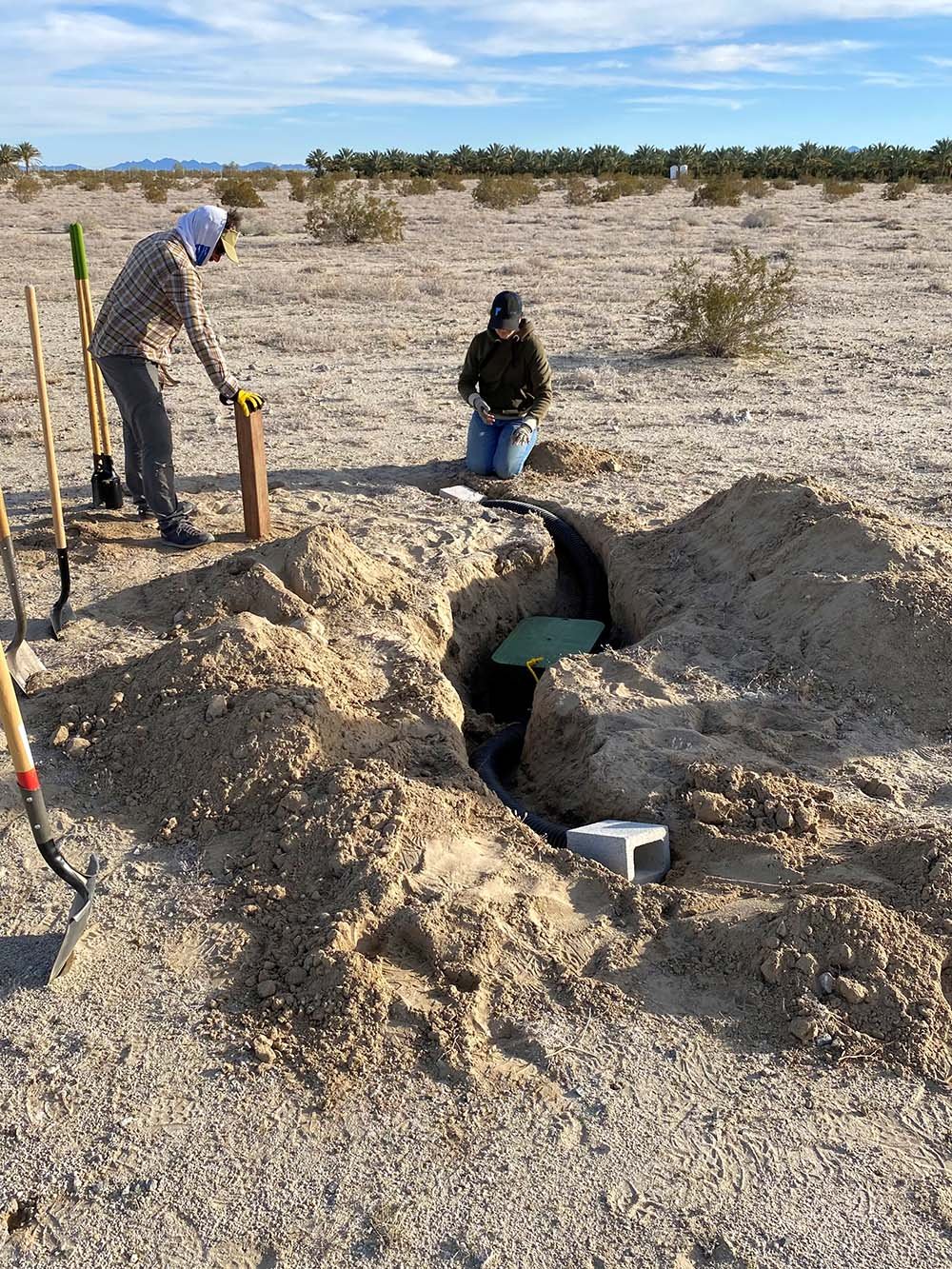 Two workers excavate a trench in a desert landscape, installing underground cables with shovels and equipment, with palm trees and mountain ranges in the background.