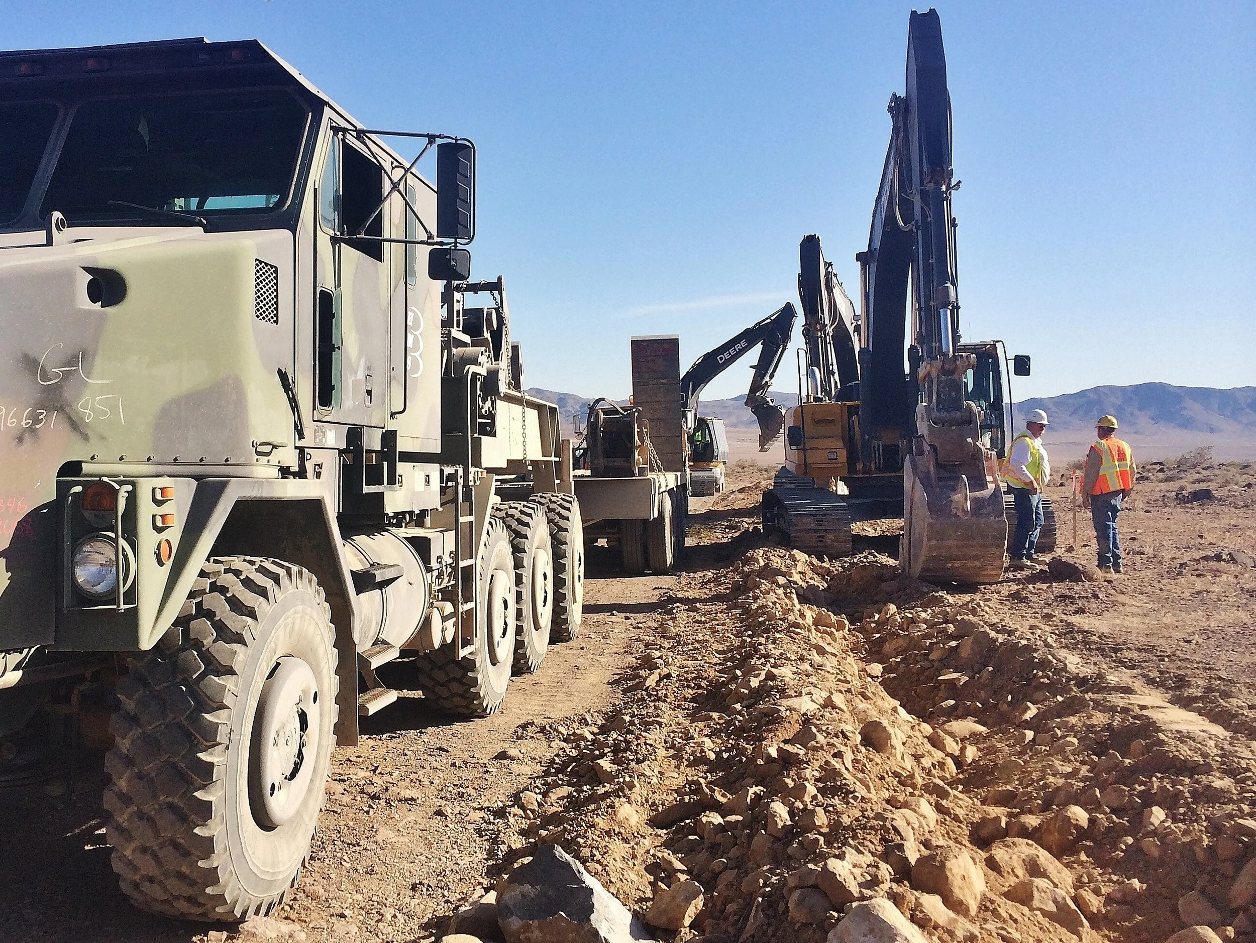 Construction site with large truck, excavator, and workers wearing safety helmets and vests, with mountains in the background under a clear blue sky.