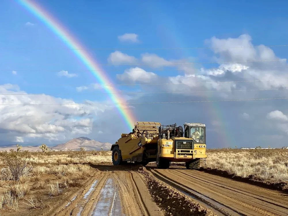 A yellow road grader on a dirt road in a desert landscape with a rainbow in the sky and clouds overhead.