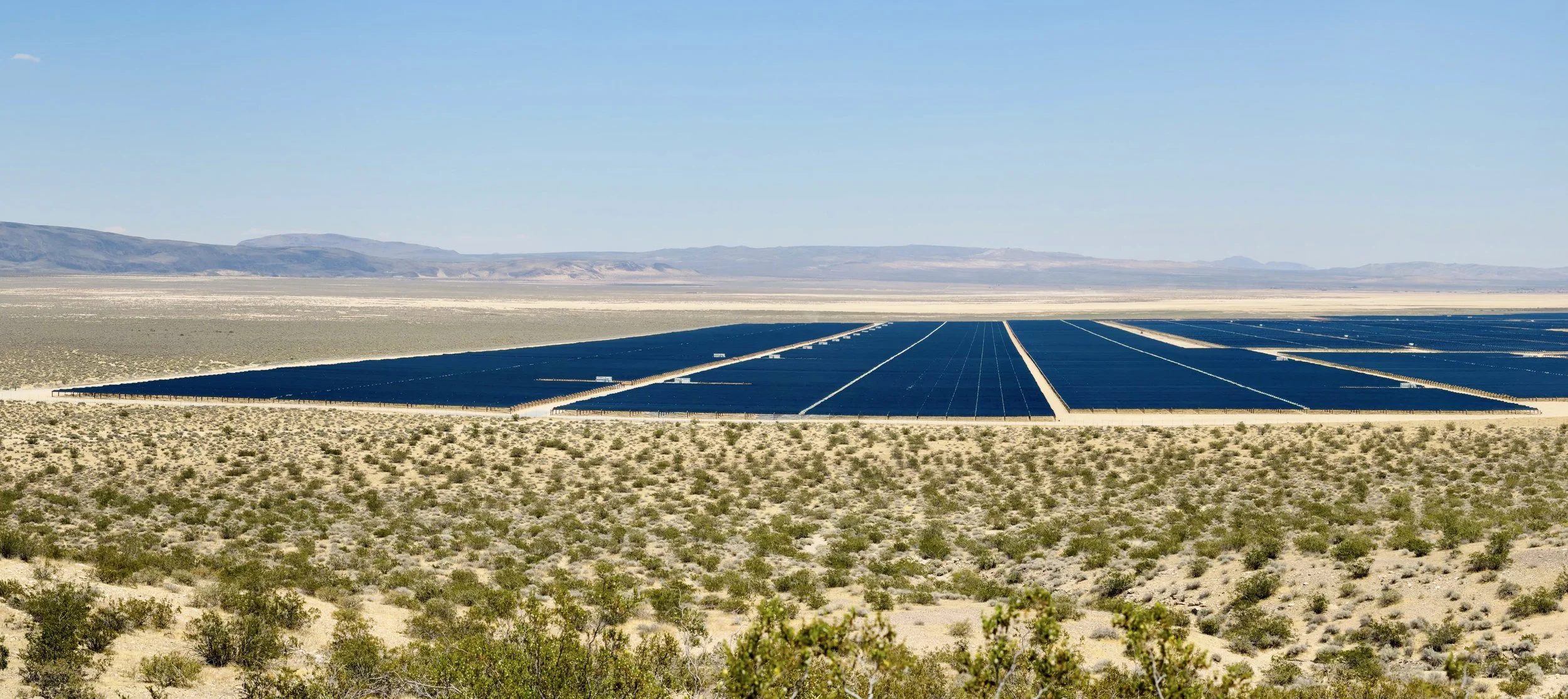 A vast desert landscape with a geothermal or solar farm containing numerous large, dark solar panels arranged in rows, with mountains in the background under a clear blue sky.