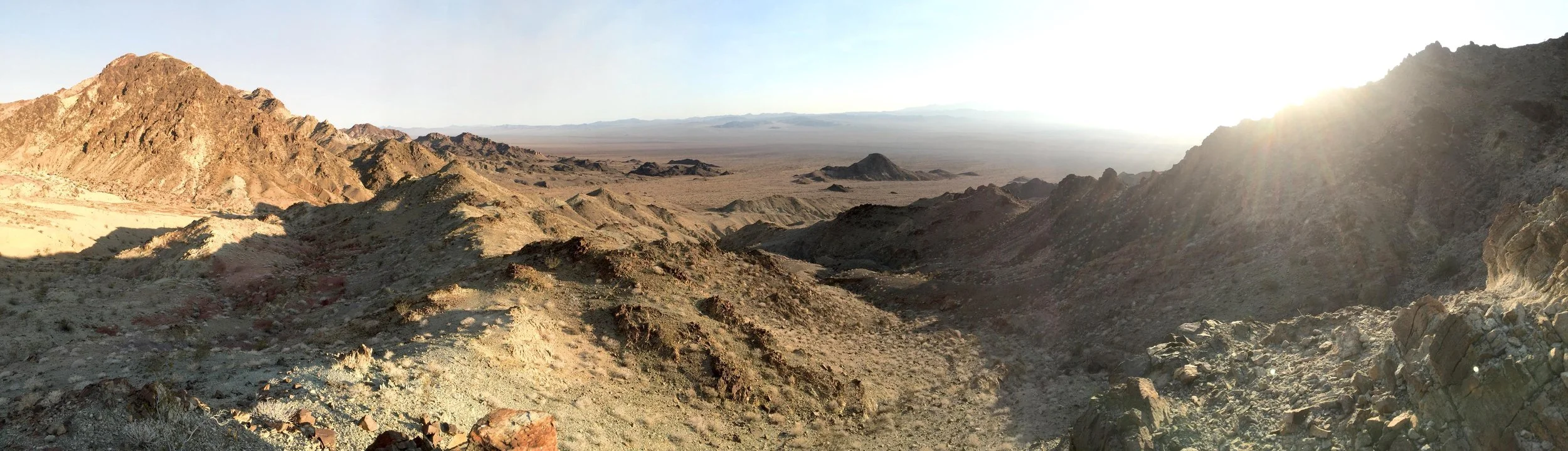 A panoramic view of a desert landscape with rocky hills and mountains under a bright sky with the sun low on the horizon.