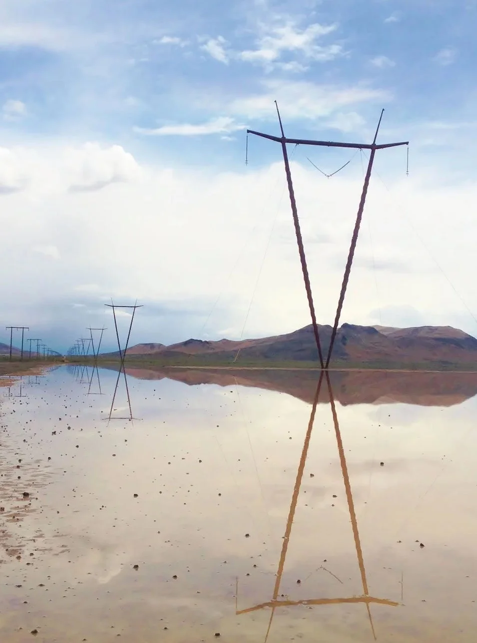 A landscape with power lines and a large puddle reflecting the sky and hills on a cloudy day.