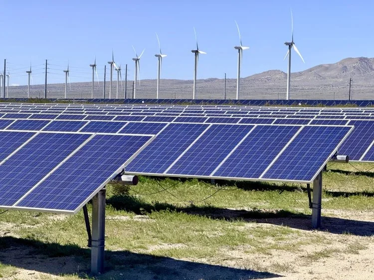 A solar farm with solar panels in the foreground and wind turbines on a mountain range in the background under a clear blue sky.