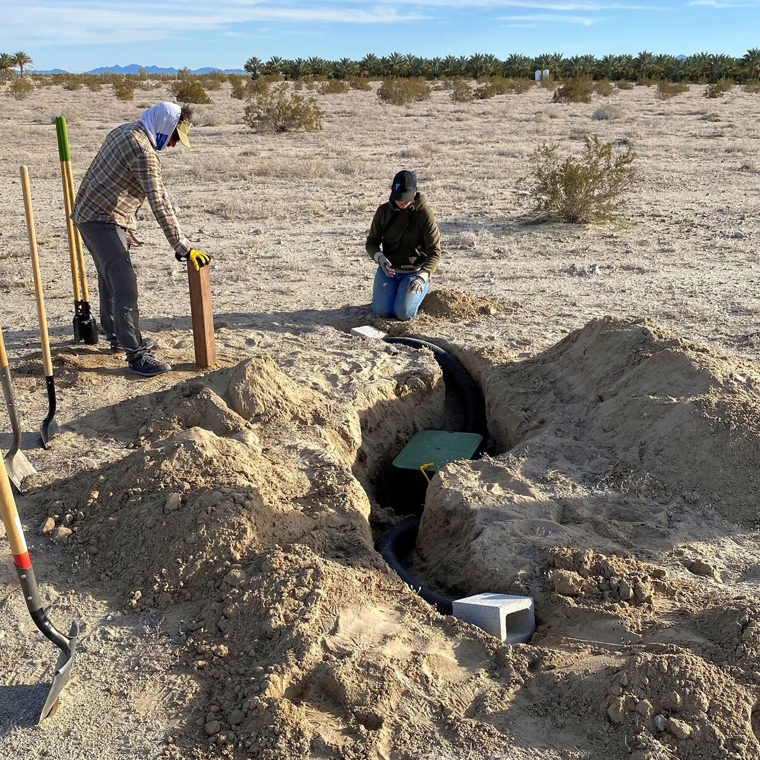 Two people working at an excavation site in a desert landscape. One person is kneeling inside a trench with a black corrugated pipe, and the other is standing nearby holding a piece of wood.