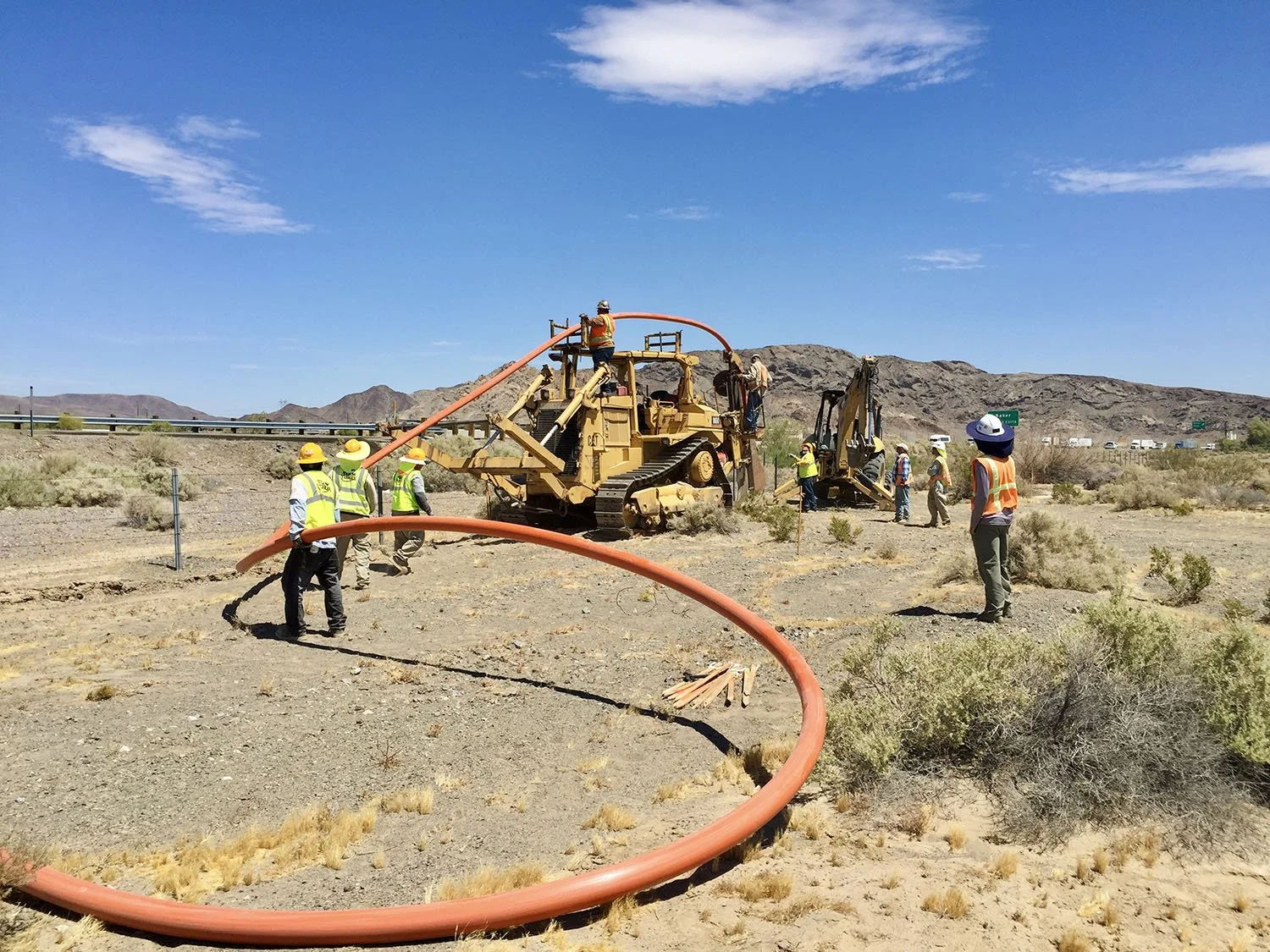 Construction workers and machinery working on a pipeline in a desert landscape under a blue sky.