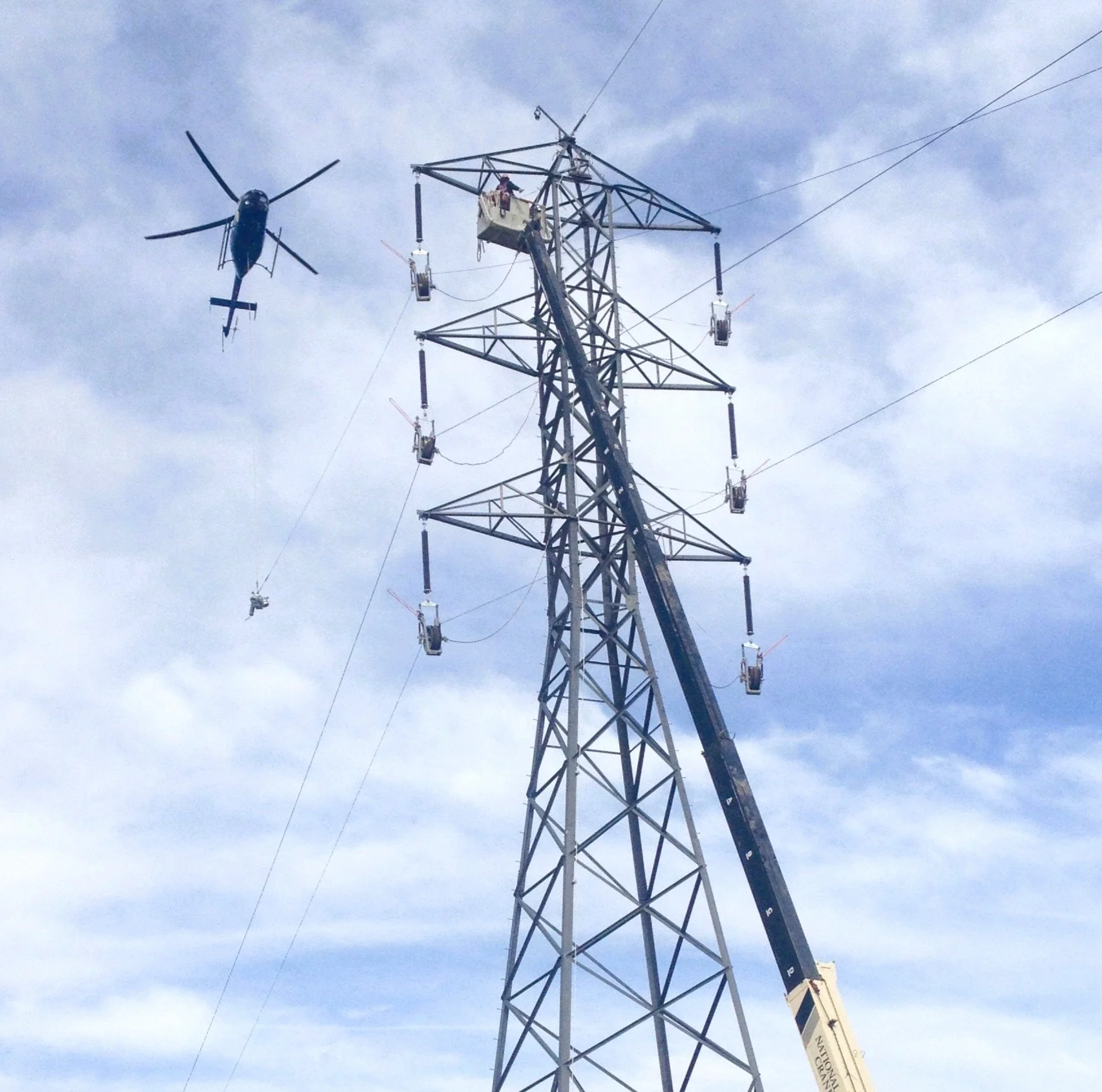 A helicopter flying near a tall electrical pylon with power lines, with a worker on a lift working on the pylon.