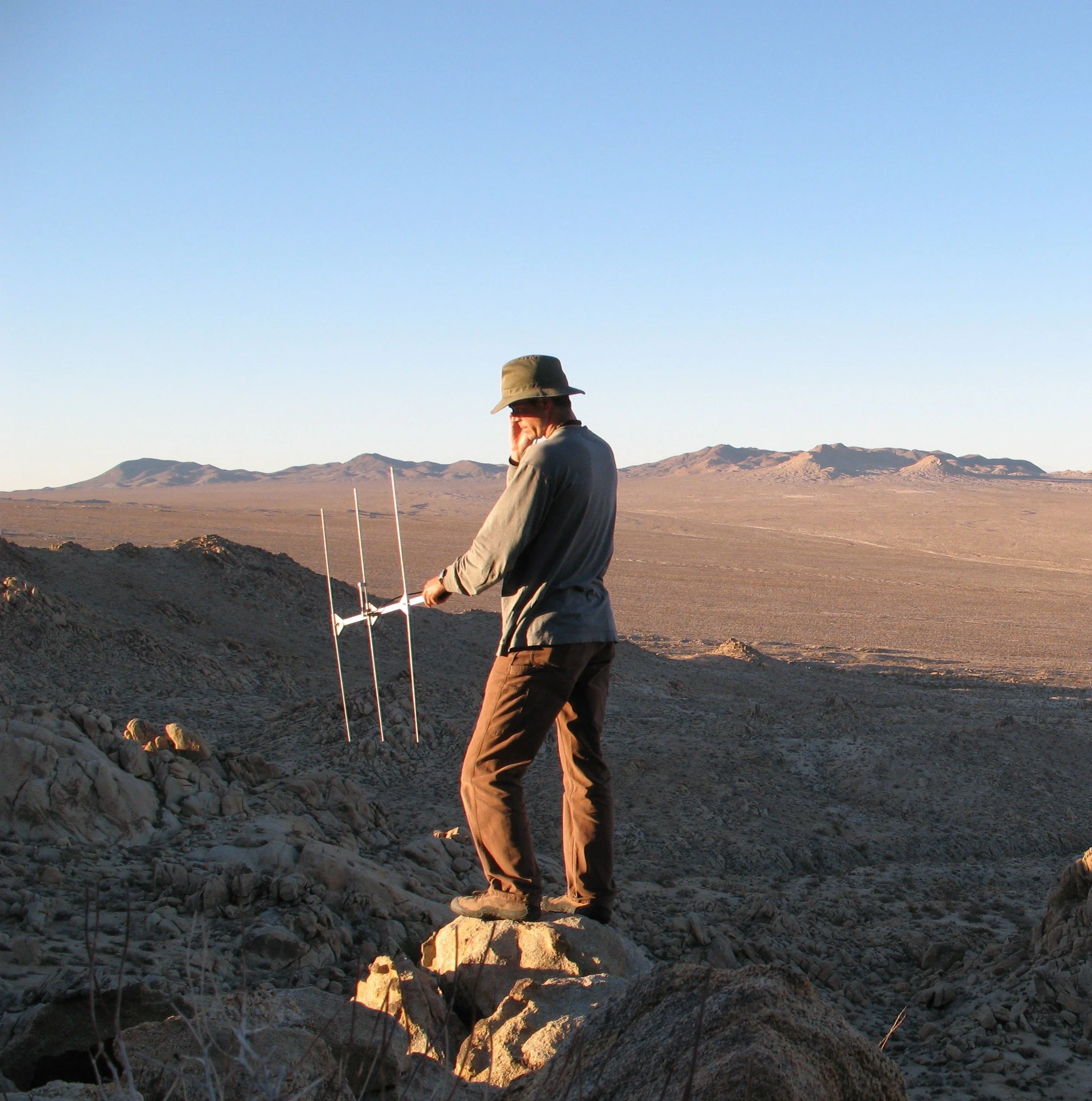 A person wearing a wide-brimmed hat and outdoor clothing standing on rocky terrain in a desert landscape with mountains in the background during sunset.