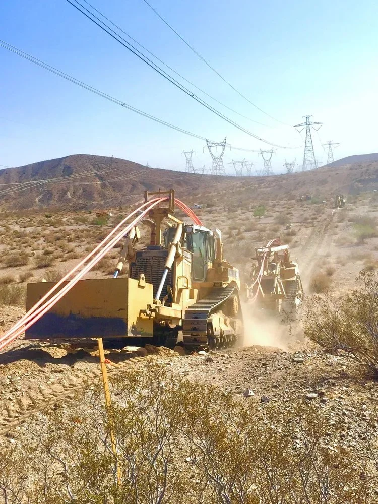 Two large construction vehicles working on a dry, desert-like hillside, with power lines and electrical towers in the background.