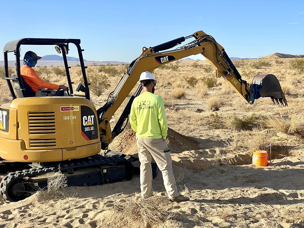 A construction worker and a person operating a CAT mini excavator digging in a desert landscape.