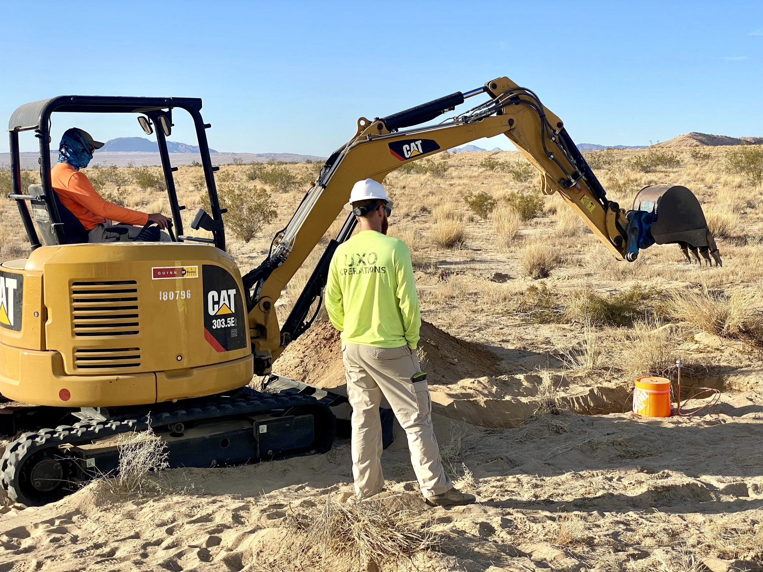 Construction workers operating a small excavator in a desert landscape. One worker is in the excavator's cab, another standing outside wearing a yellow safety vest and white hard hat. The background features sparse shrubbery and distant mountains under a clear blue sky.