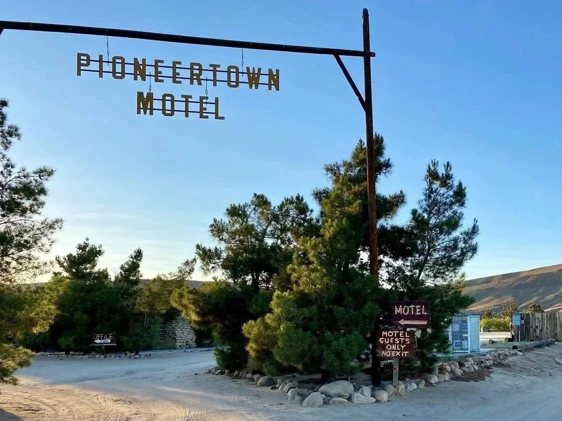 Sign for Pioneertown Motel hanging from a metal arm, surrounded by trees and gravel ground, with additional signs indicating office and motel guest parking with no exit, in a desert landscape with hills in the background.