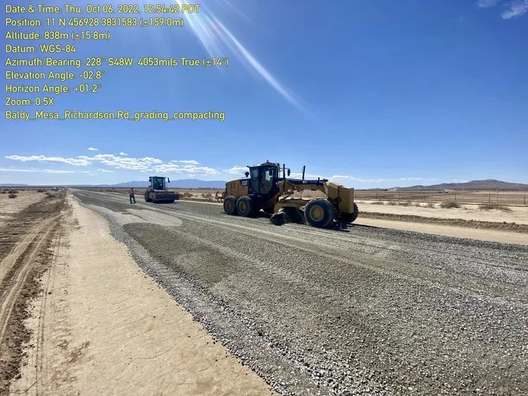 A road construction site in a desert landscape with gravel paving equipment and workers, under a blue sky with some clouds.