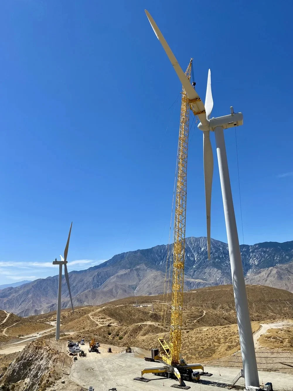 Construction crane installing a wind turbine on a hill with mountains in the background, wind turbines, and a clear blue sky.