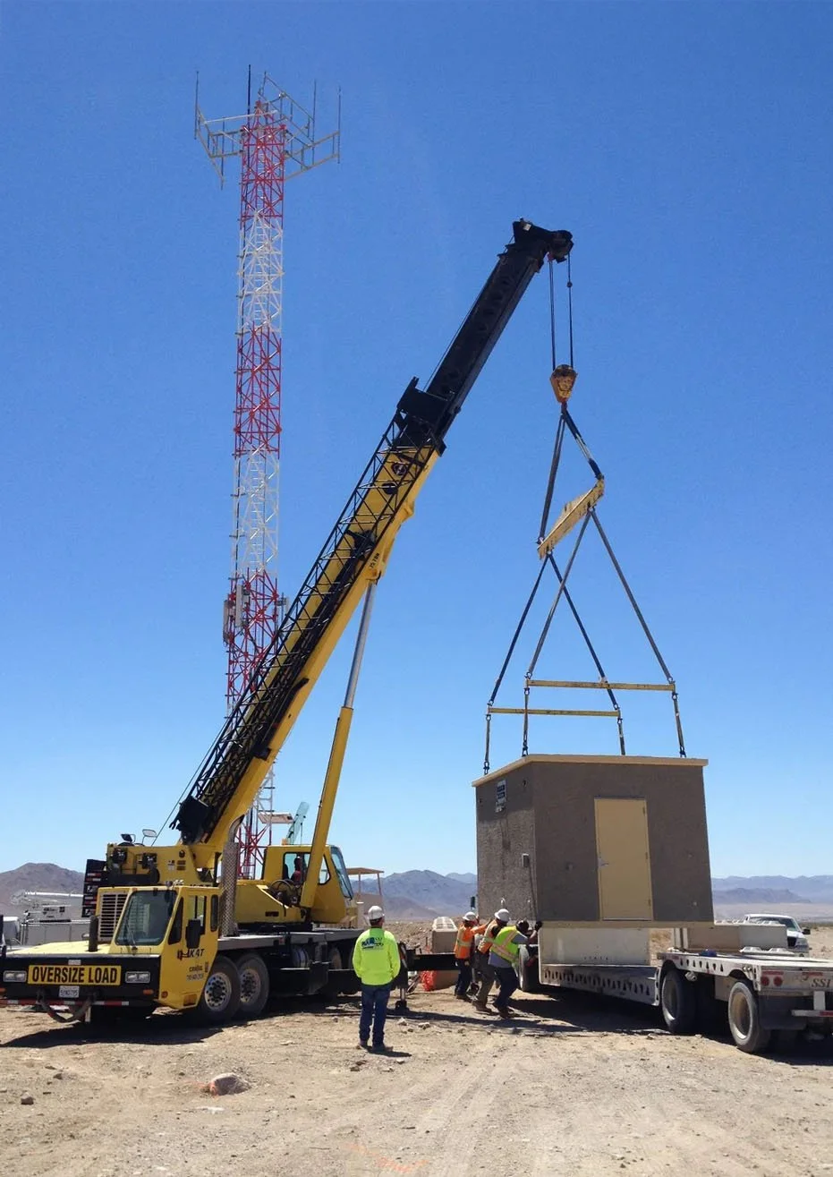 A construction site in a desert setting shows a large yellow crane lifting a concrete building module onto a flatbed truck, with workers in safety vests and helmets guiding the process.