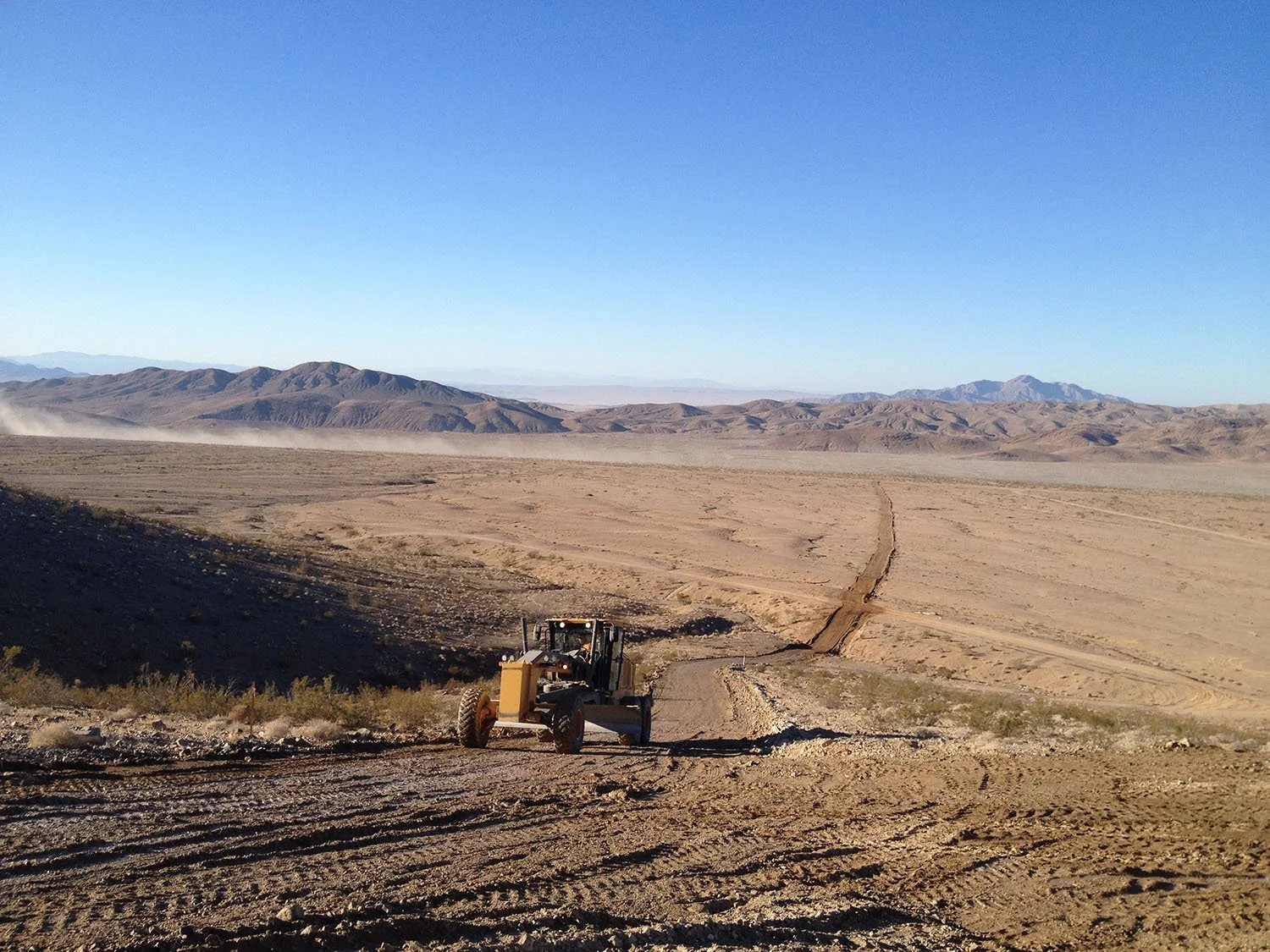 Construction vehicle on a dirt road in a desert with mountains in the background.