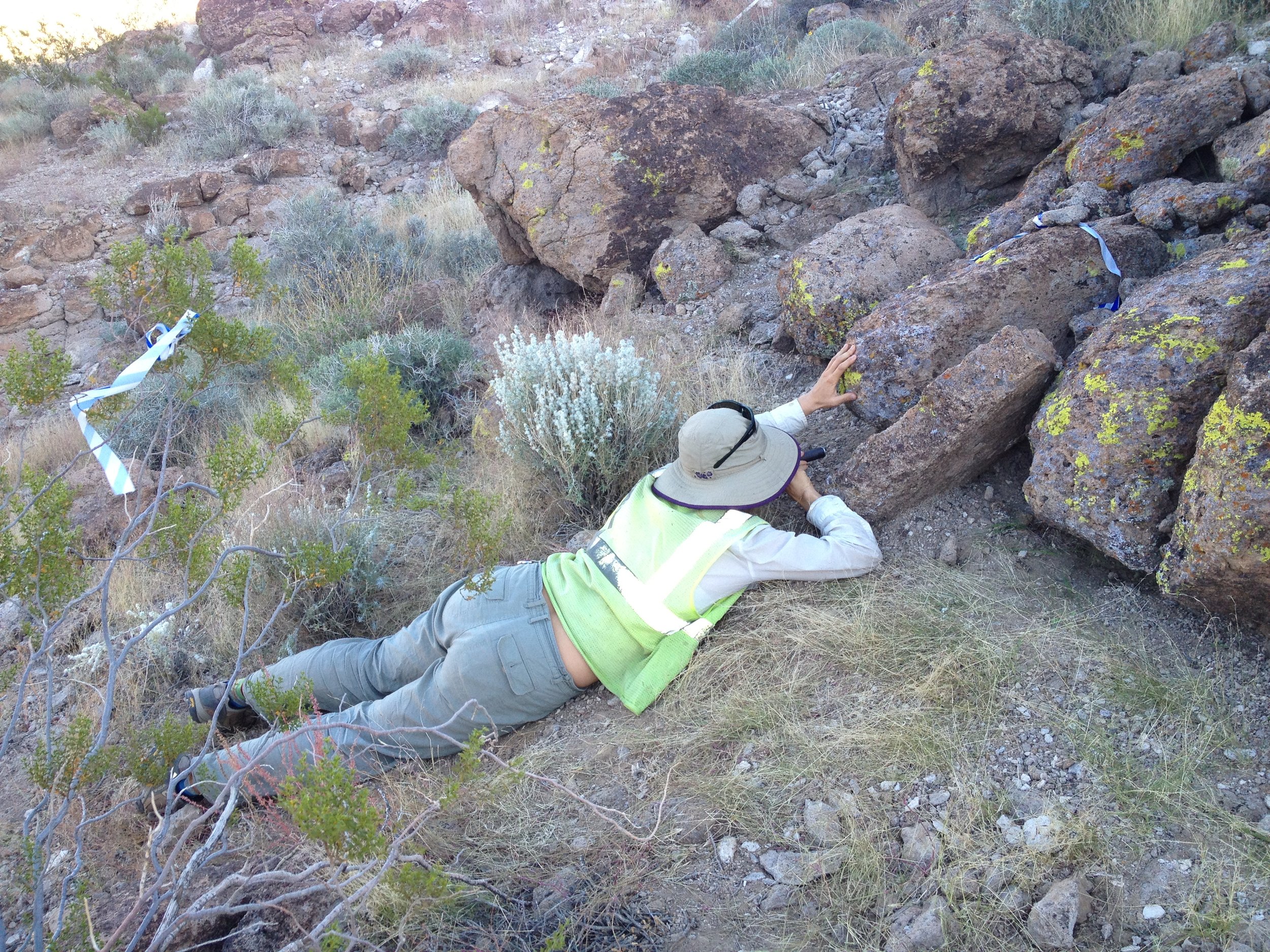 A person lying on the ground in a rocky, desert-like terrain, reaching out to touch a large boulder.