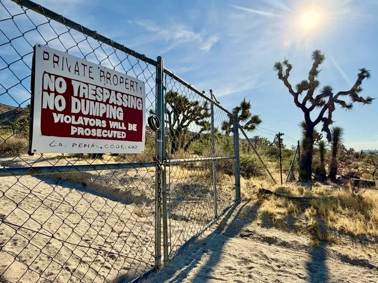 A chain-link fence with a sign that reads 'Private Property. No Trespassing. No Dumping. Violators will be prosecuted.' in a desert landscape with a Joshua tree and a bright sun in the sky.