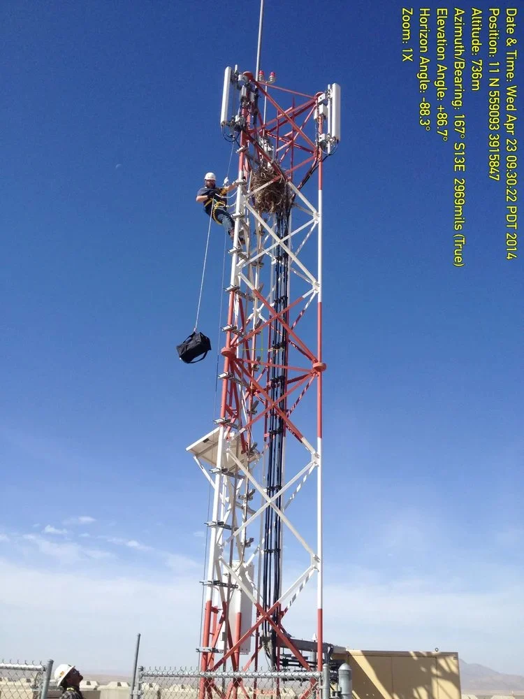 A technician working on a large telecommunications tower during daytime, with clear sky and distant mountains, wearing safety gear and climbing the metal structure.