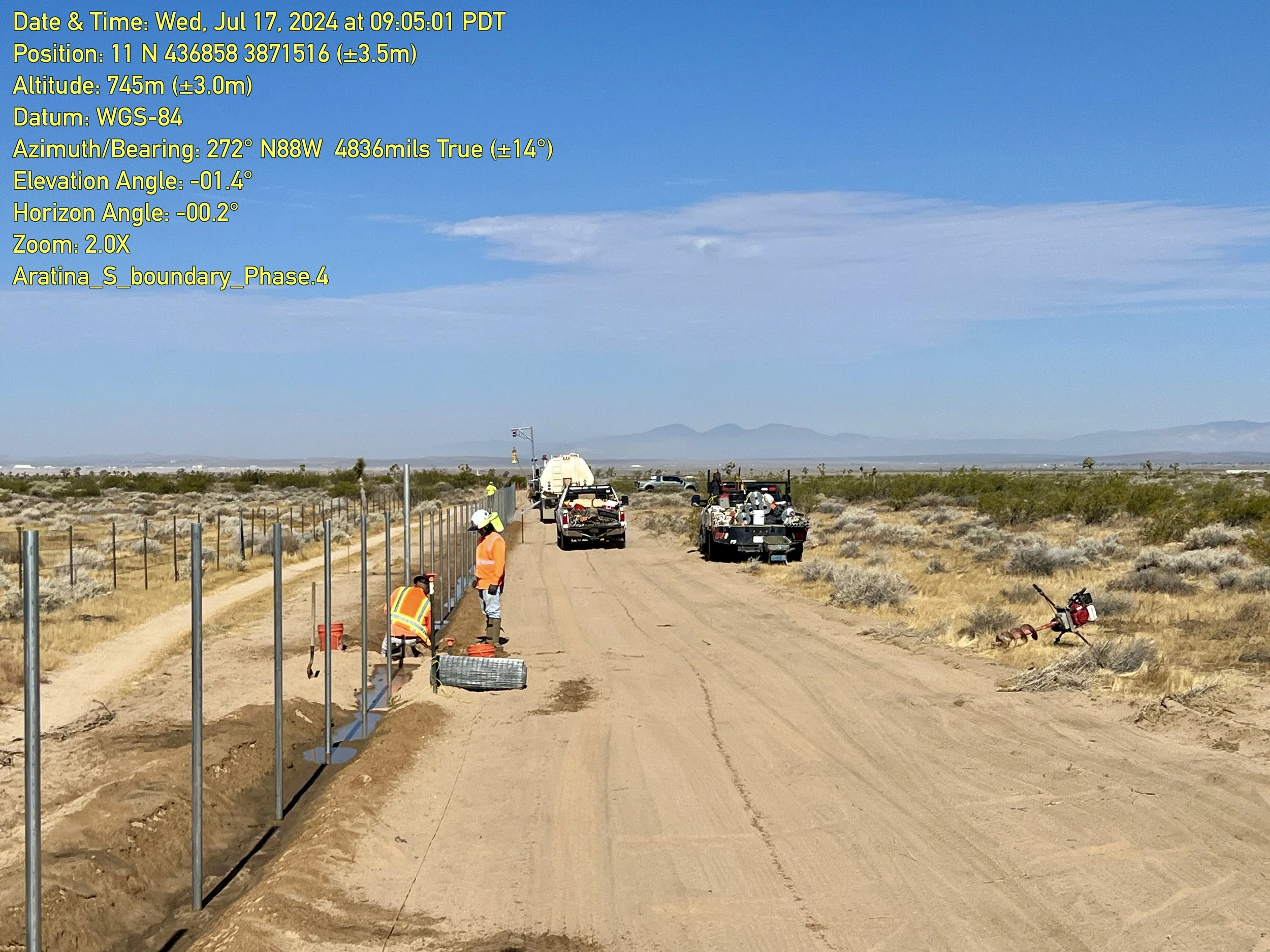 Construction workers and equipment installing fencing along a dirt road in a desert landscape, with temporary water pipes visible on the ground and mountains in the distance.