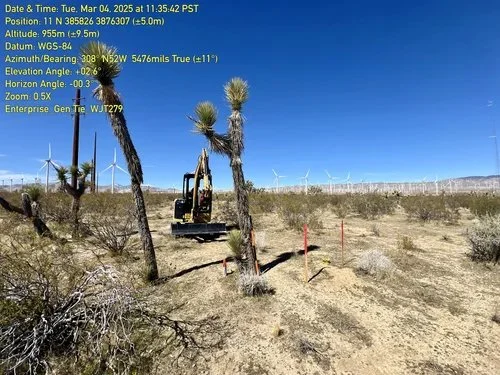Construction equipment working among desert vegetation with Joshua trees, wind turbines in the background, under a clear blue sky.