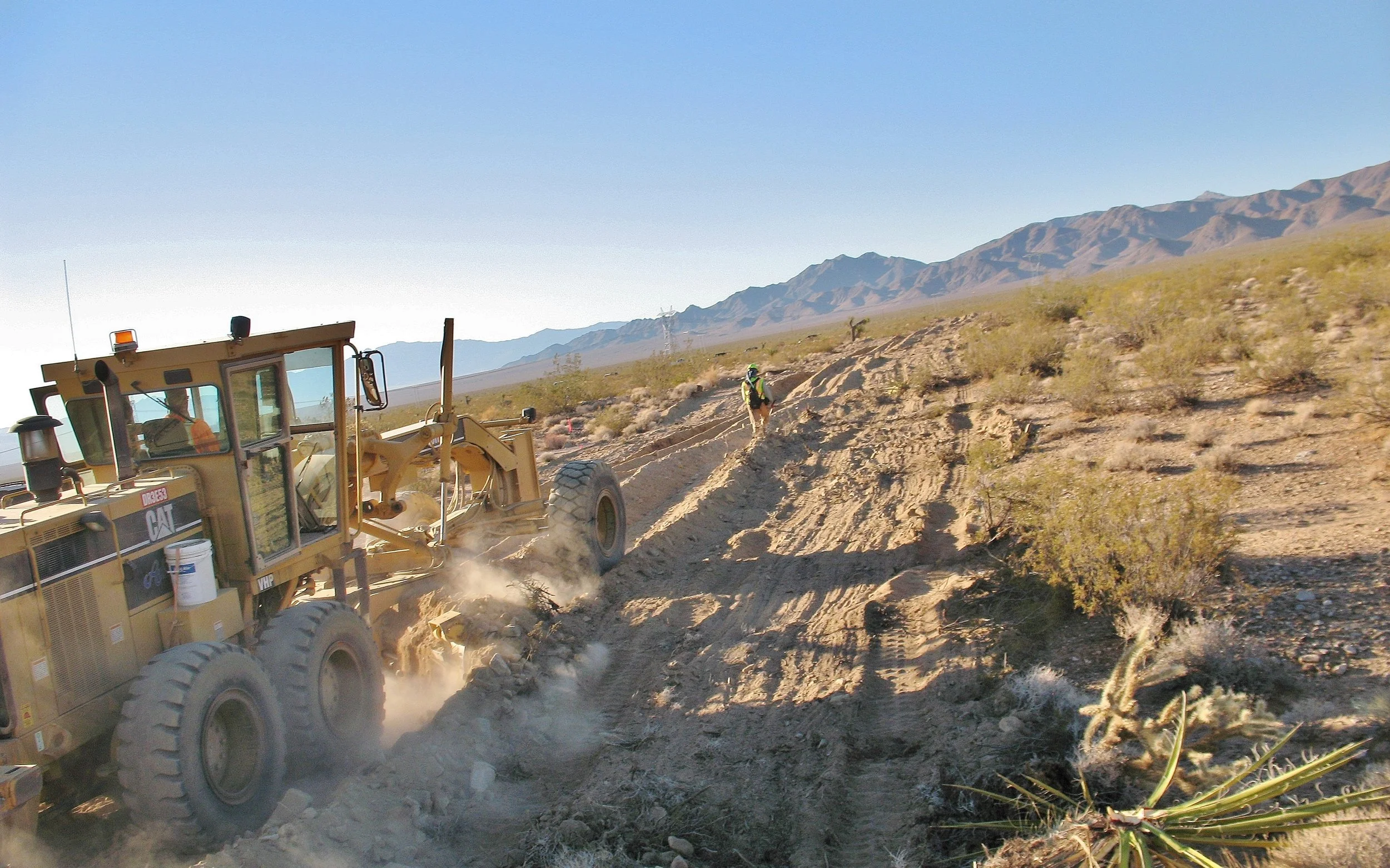 A construction vehicle working on a dirt trail in a desert landscape with mountains in the background, and a person walking along the path.