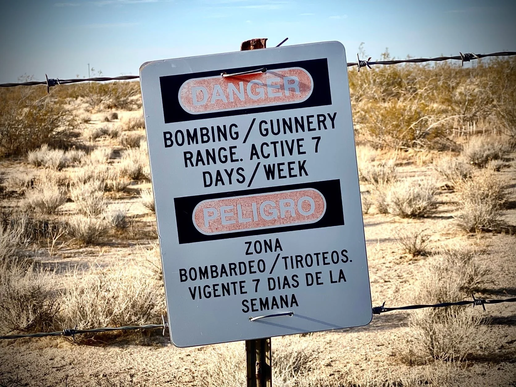 A warning sign in a desert area warning of danger from a bombing range, with active shooting days noted as 7 days a week, written in both English and Spanish, surrounded by dry bushes and barbed wire.