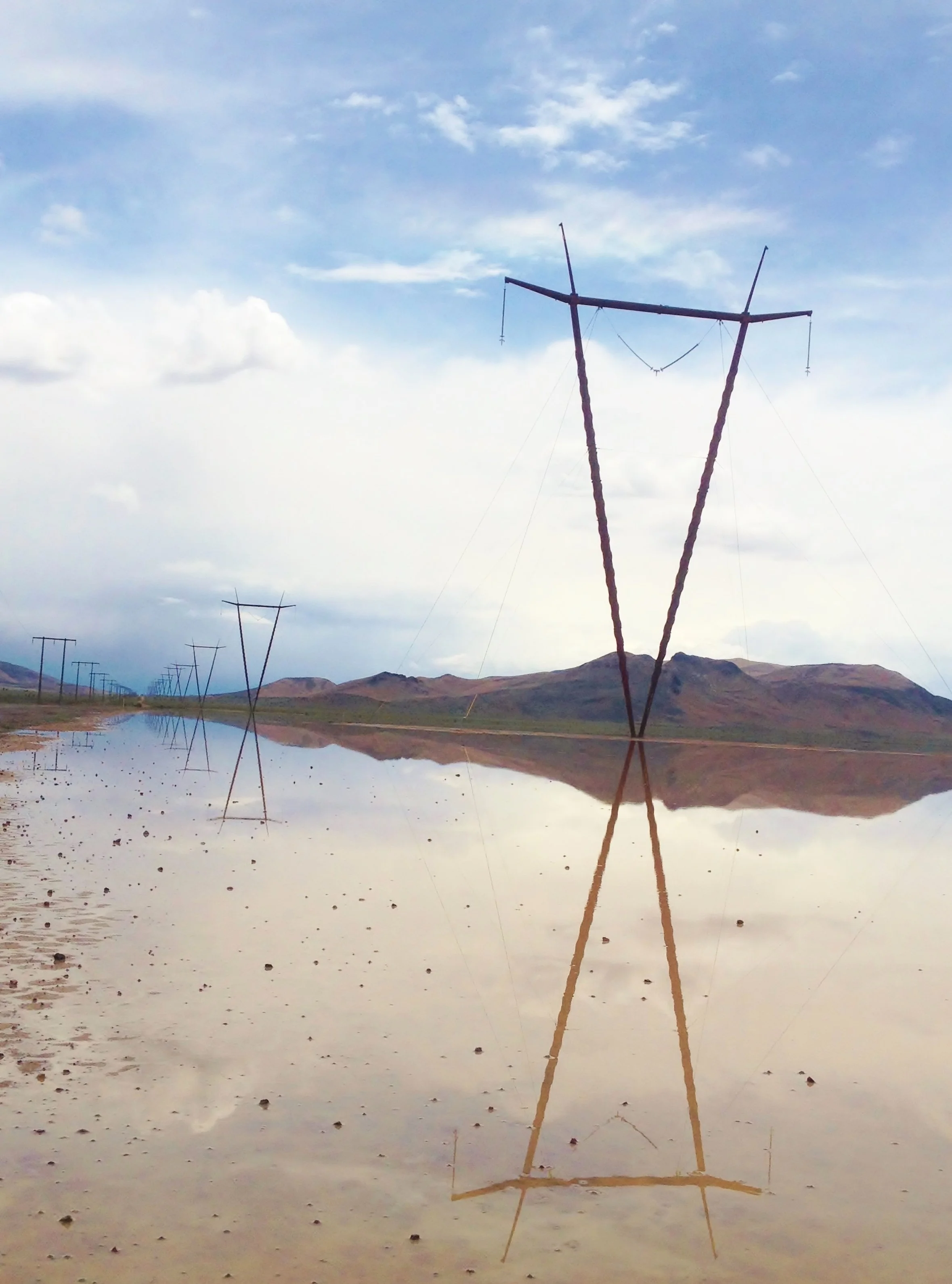 Reflection of a tall, wooden, Y-shaped utility pole with wires hanging from it in a body of water with mountains and a partly cloudy sky in the background.