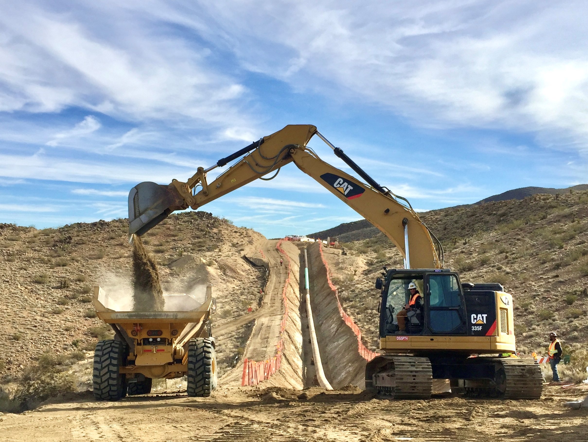 Construction site with a large yellow Caterpillar excavator and a smaller bulldozer working on a dirt road in a hilly, desert landscape under a partly cloudy sky.