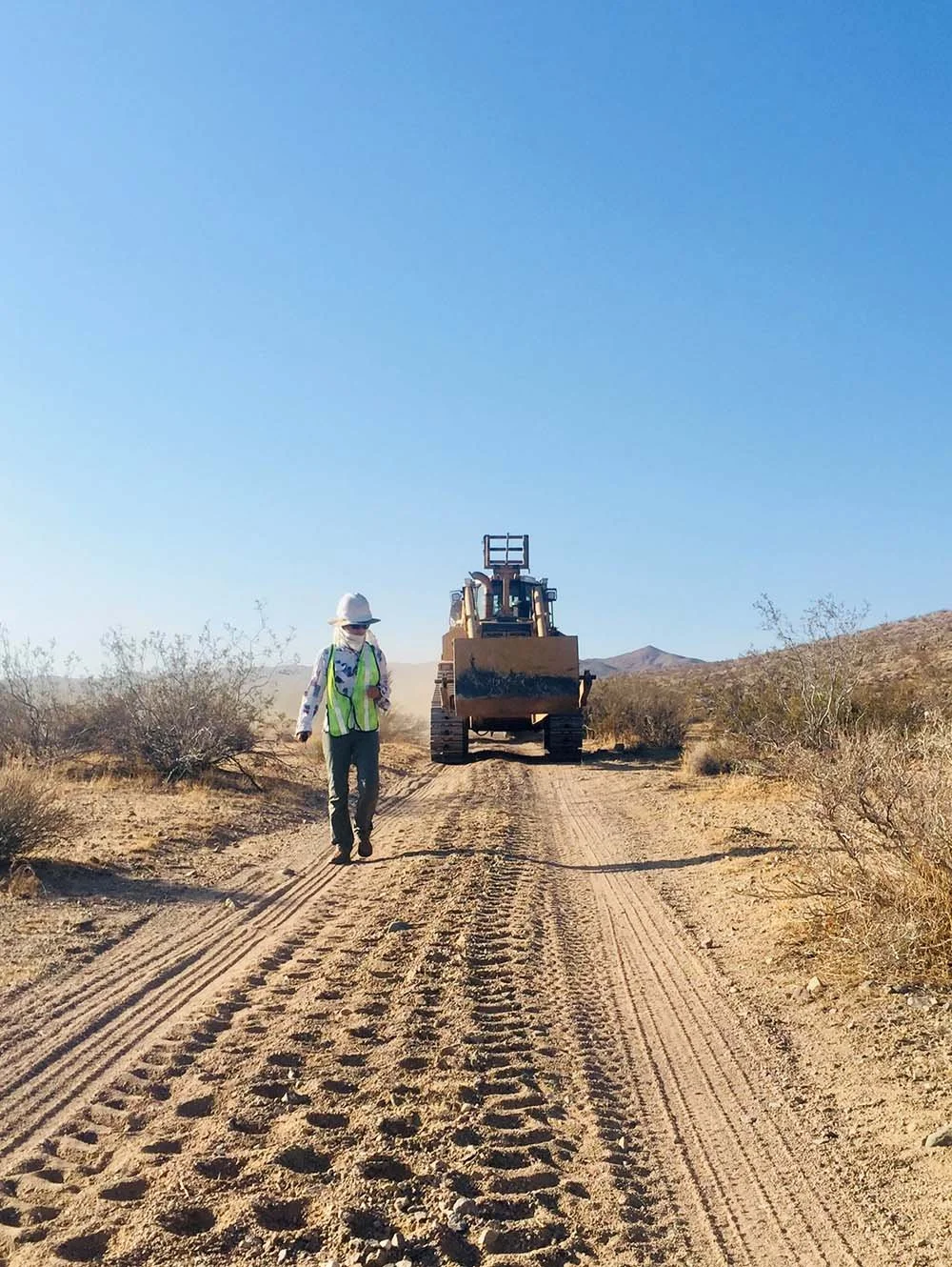 A construction worker walking on a dirt road in a desert area with a bulldozer behind him under a clear blue sky.