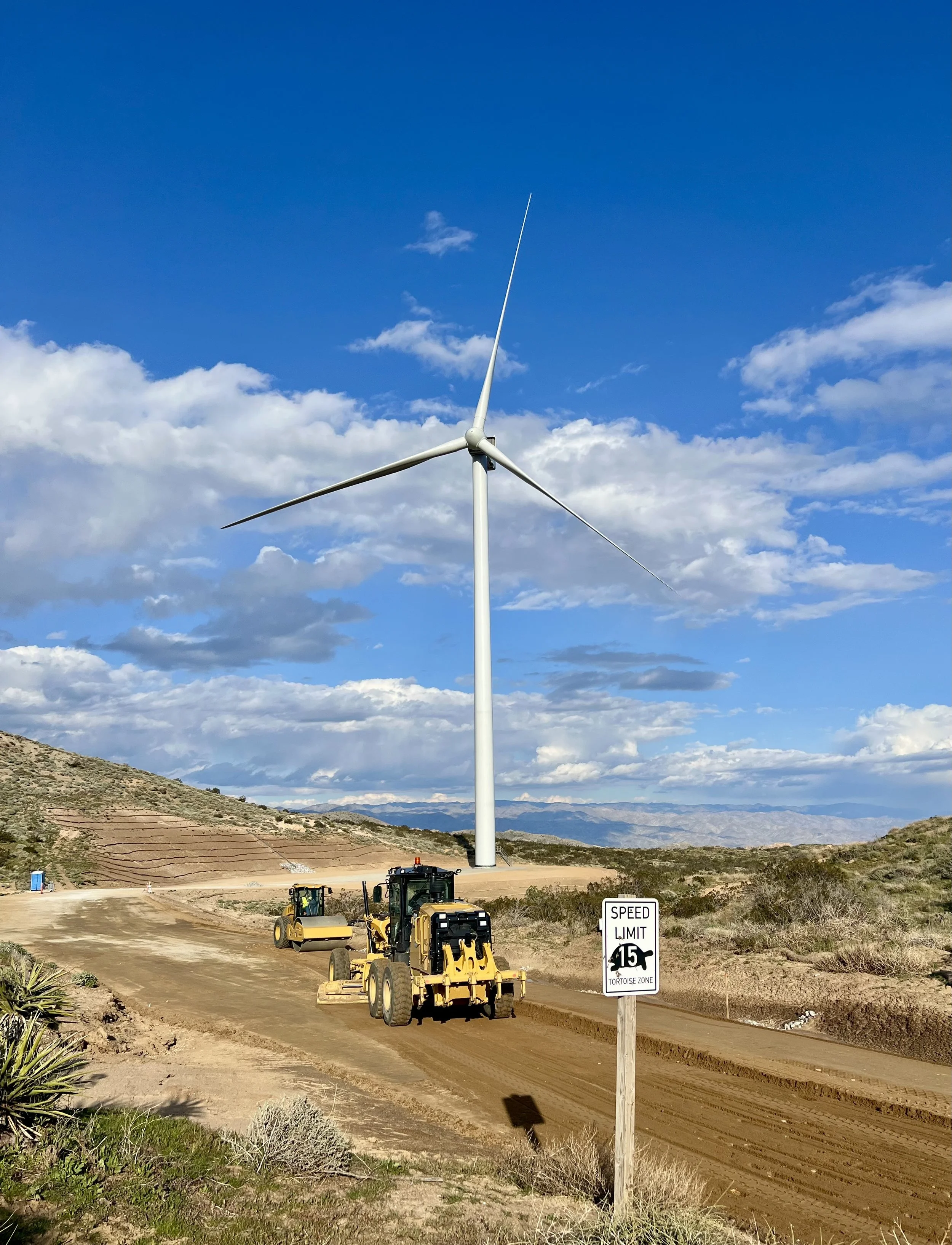 A wind turbine under construction in a mountainous area with construction vehicles and a speed limit sign that says 15 miles per hour.
