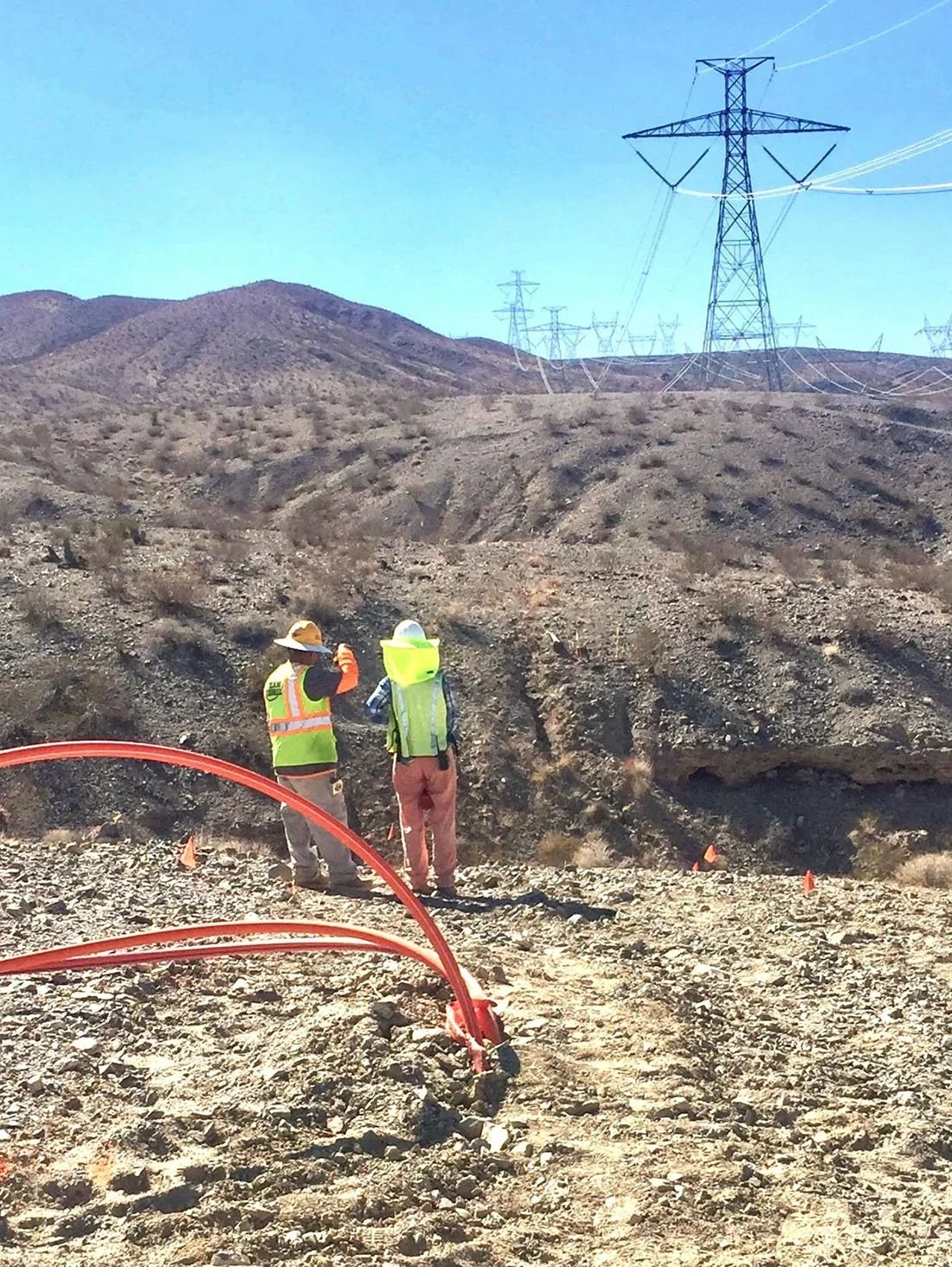 Two workers in safety gear stand on a dirt construction site in a desert landscape, with large electrical transmission towers in the background.