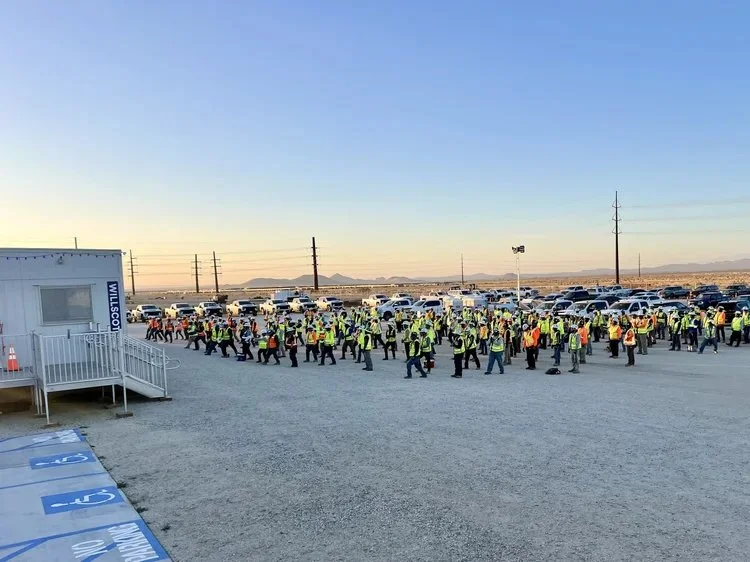 Large group of people wearing yellow safety vests walking in an outdoor parking lot at sunset.