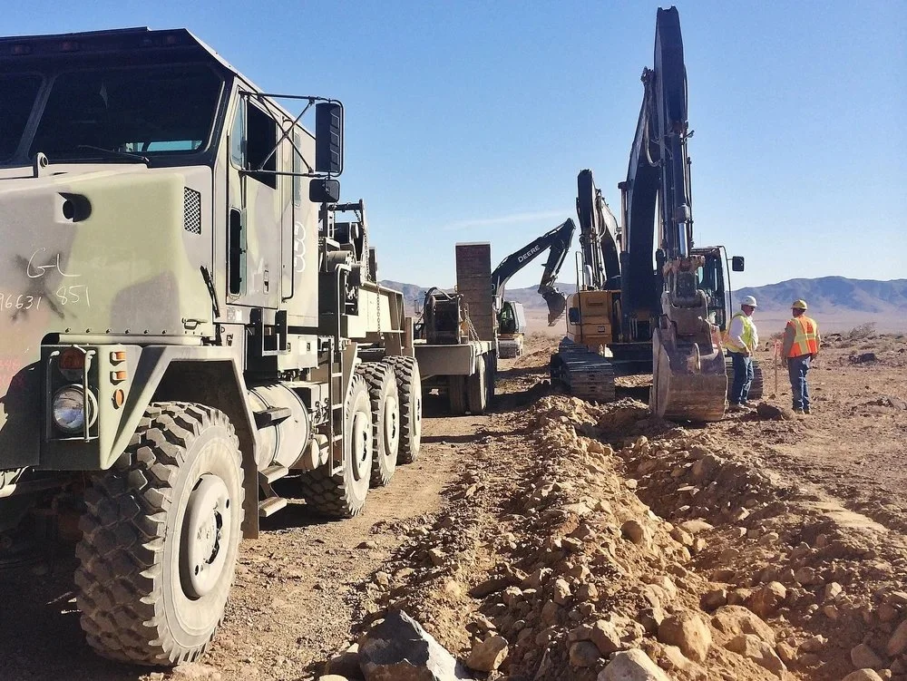 Construction site in a desert with large trucks, excavators, and workers wearing safety vests and helmets, digging into the ground.