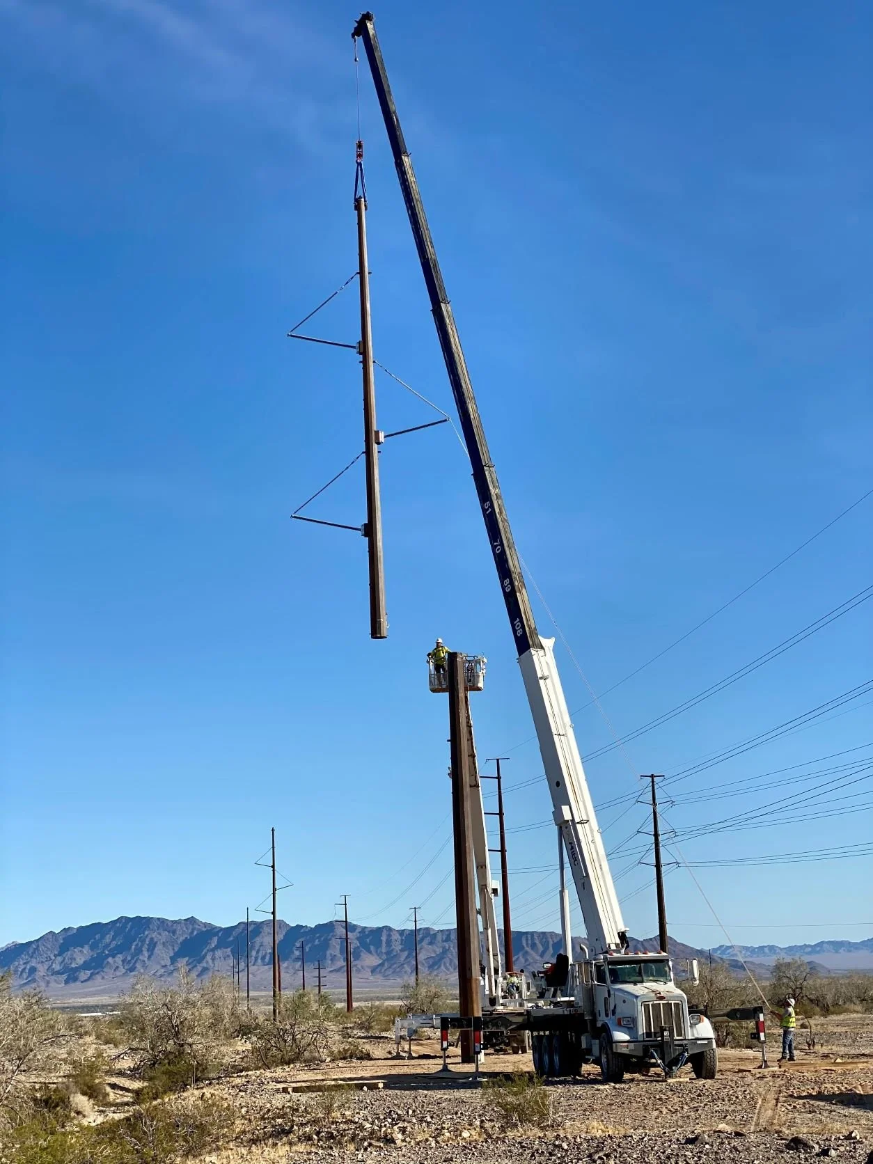 A crane lifting a utility pole in a desert area with mountains in the background, with workers present.