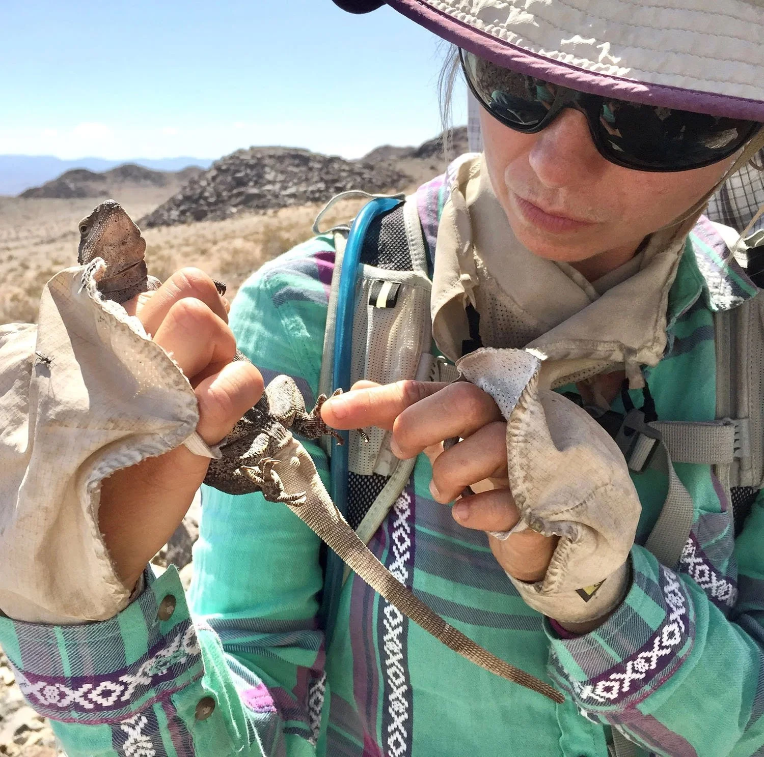 A woman wearing a sun hat, sunglasses, and a colorful jacket is holding a small lizard in her hands in a desert landscape with rocky hills in the background.