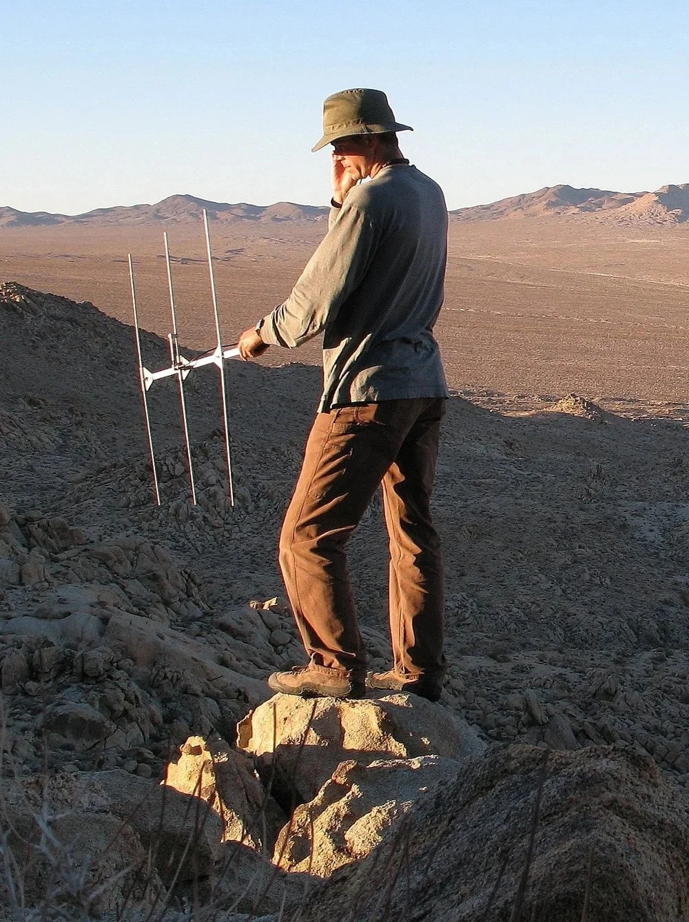 Person in outdoor gear standing on rock in a desert landscape, using a portable radio antenna for outdoor research or communication.