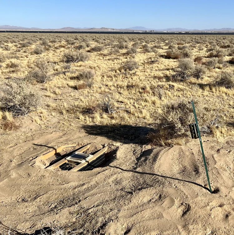 An open manhole cover in a desert landscape with sparse bushes and distant mountains under a clear blue sky, with a long metal pole propped against the ground nearby.