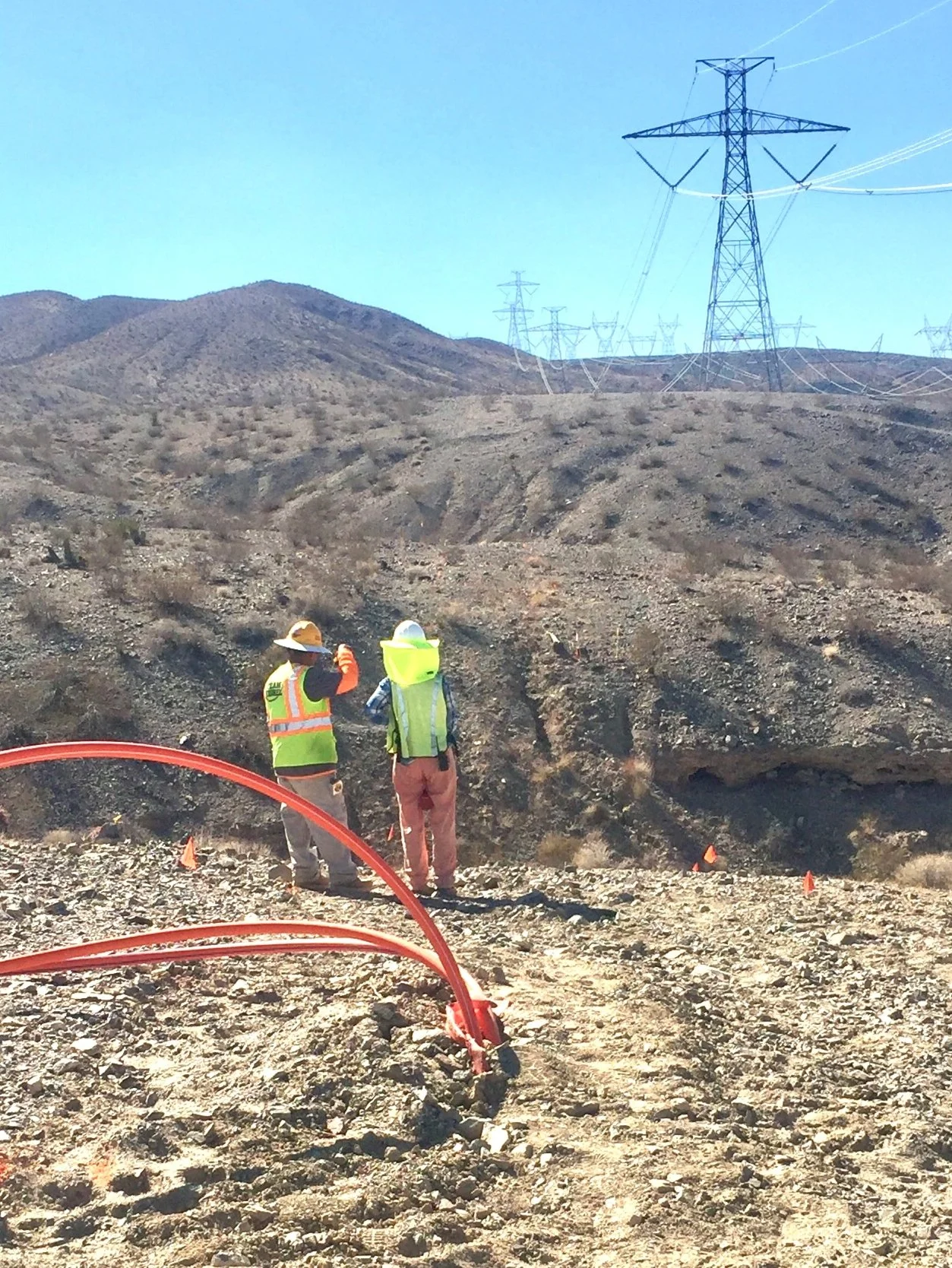 Two construction workers in safety vests and helmets standing on rocky terrain with a mountain backdrop; power lines and pylons in the distance.