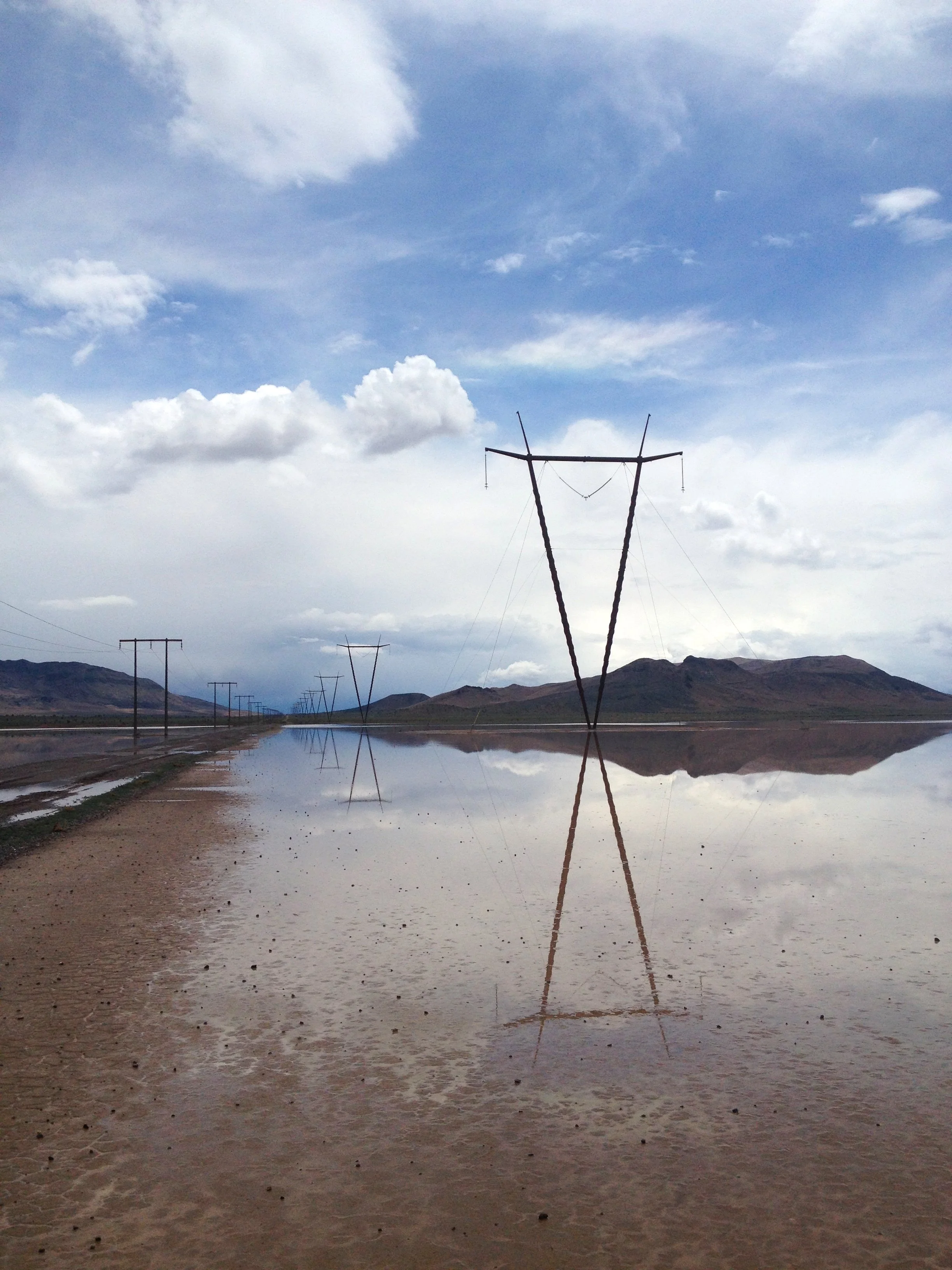 Reflection of electricity pylons on a body of water with a mountainous landscape in the background under a partly cloudy sky.