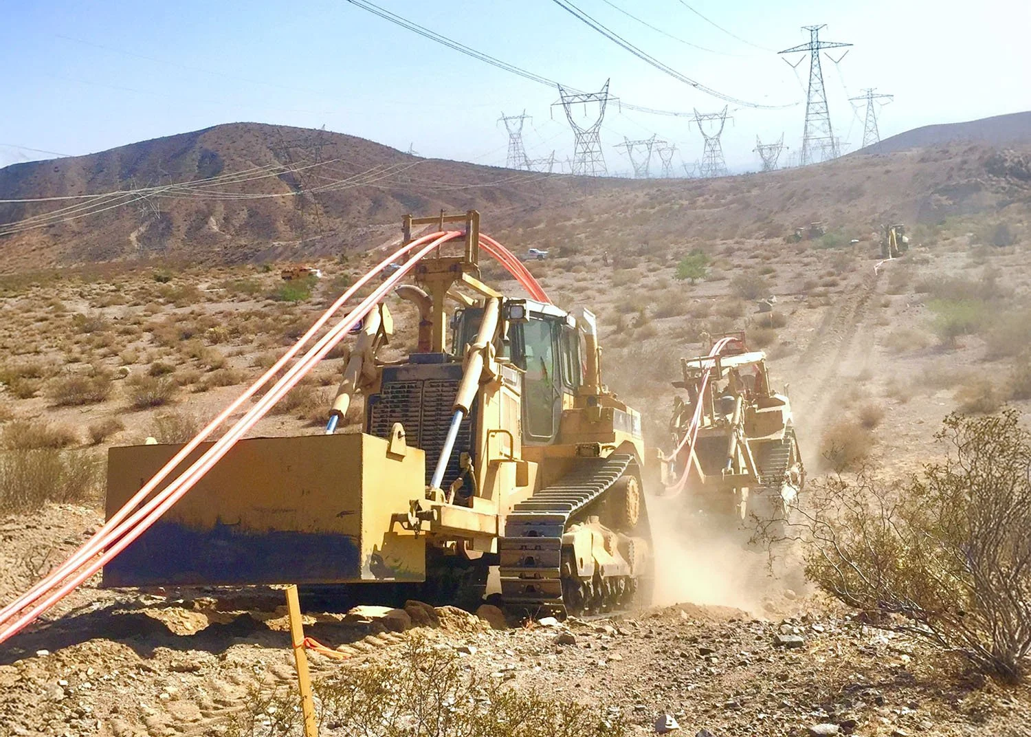 Construction site in a desert landscape with large machinery drilling for pipelines, with power lines and hills in the background.