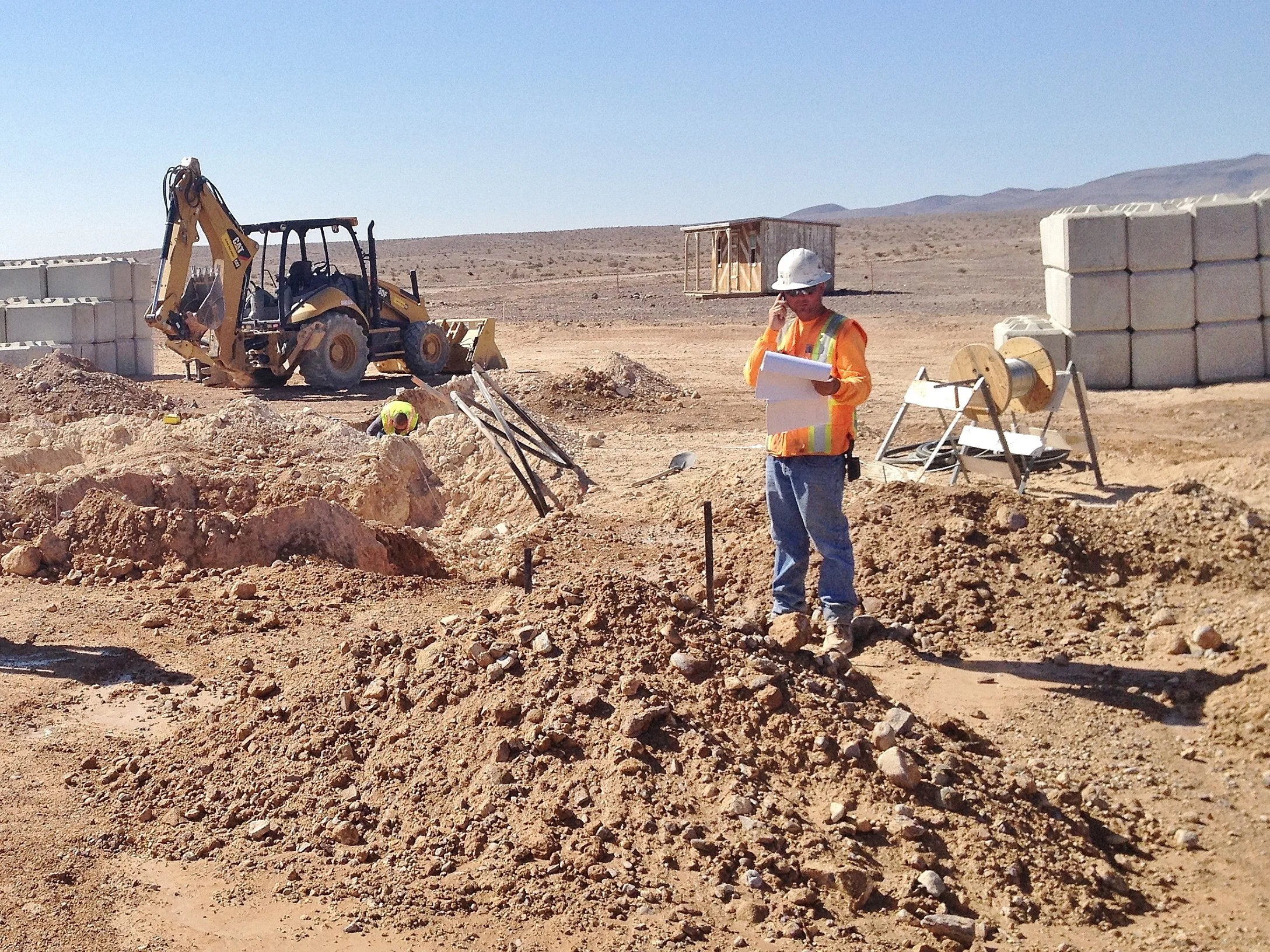 Construction site in a desert landscape with a backhoe loader, a construction worker with a walkie-talkie, and stacks of concrete blocks.