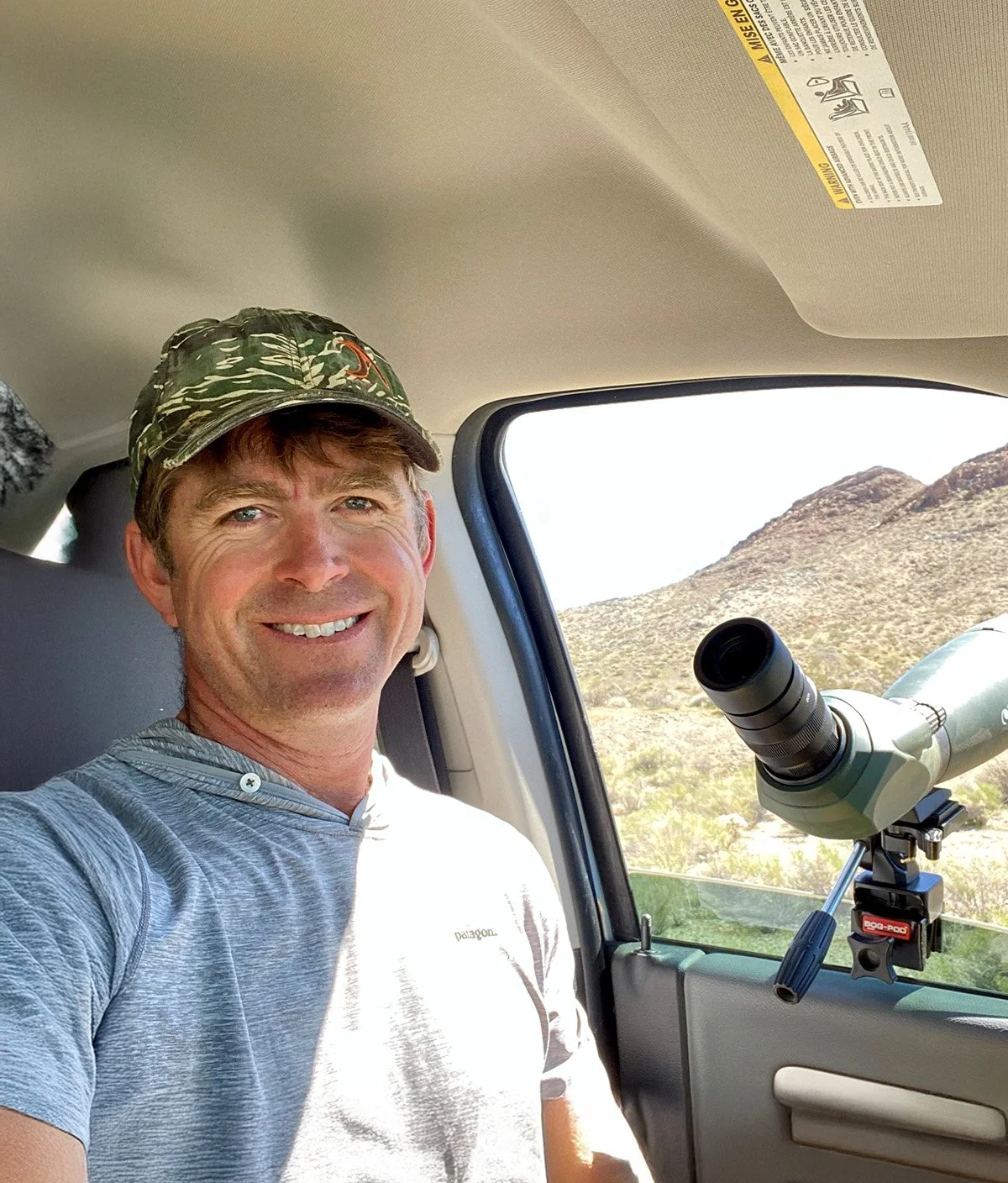 Man smiling inside a vehicle with a telescope mounted on the window, desert landscape with hills outside