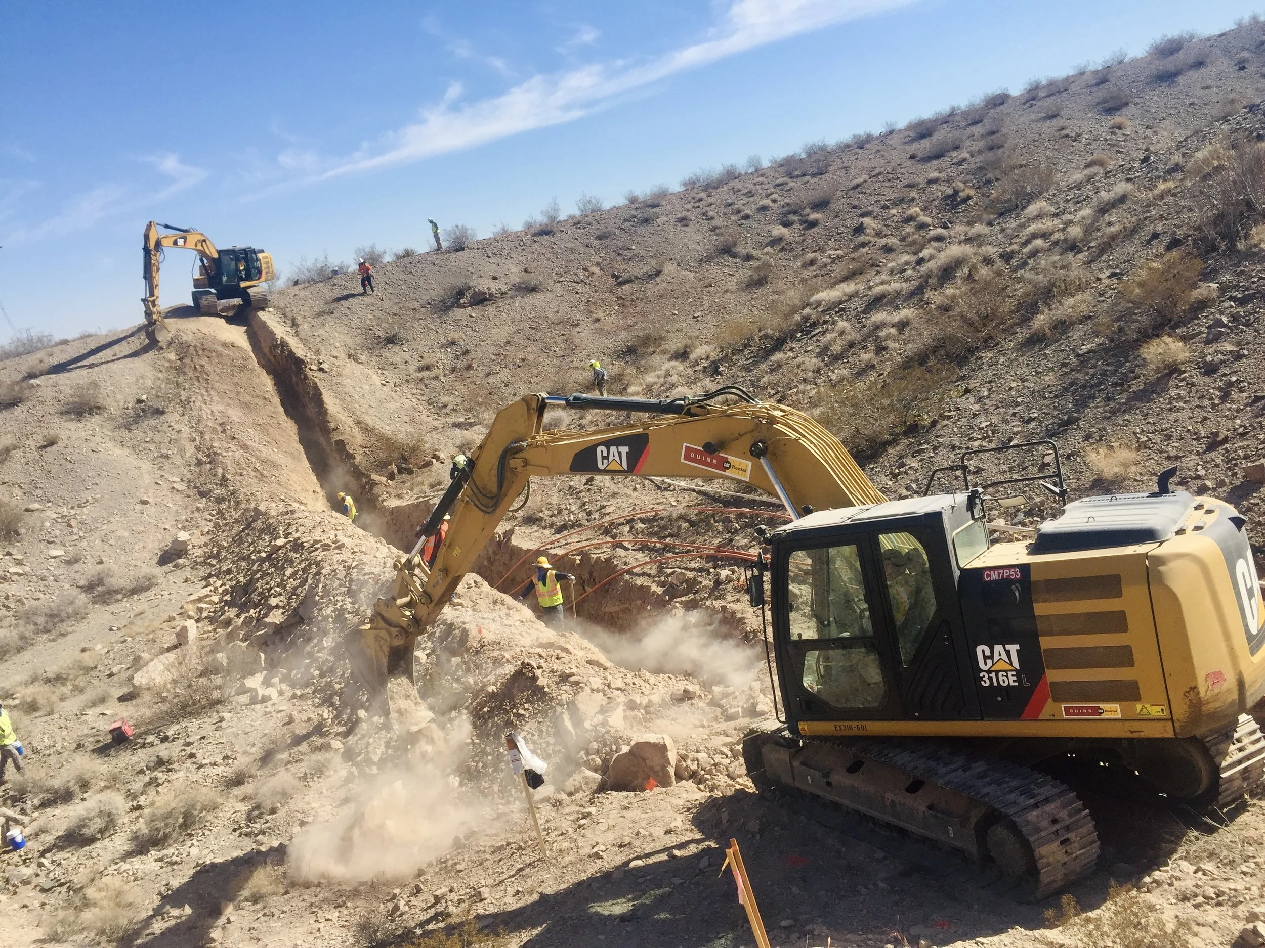 Construction site in a desert-like area with a yellow excavator digging into rocky terrain, several workers in safety gear are working around the site, and a clear blue sky overhead.