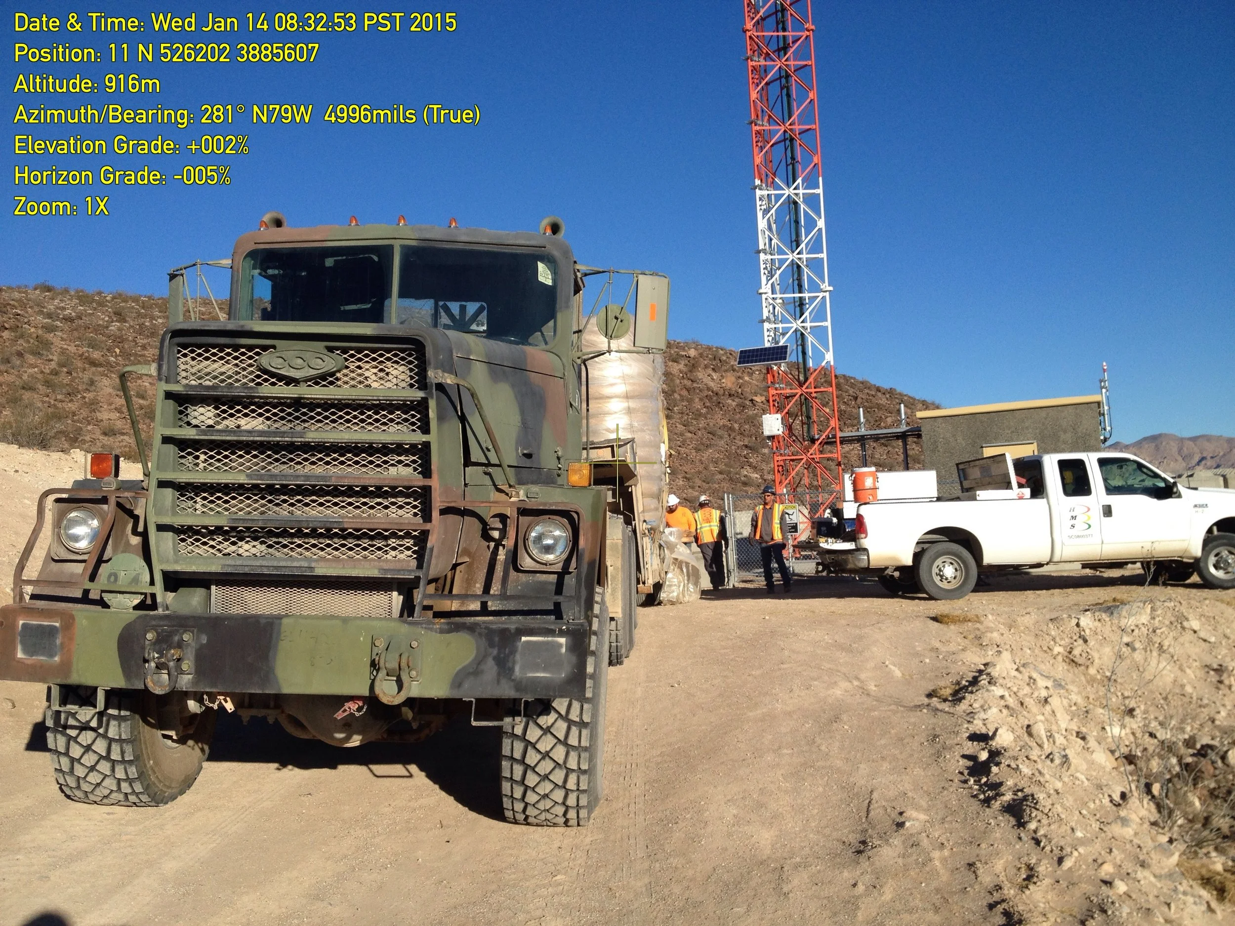 A green military truck parked on a dirt area with a large red and white radio tower and people in construction vests working nearby in the background. The sky is clear and blue.