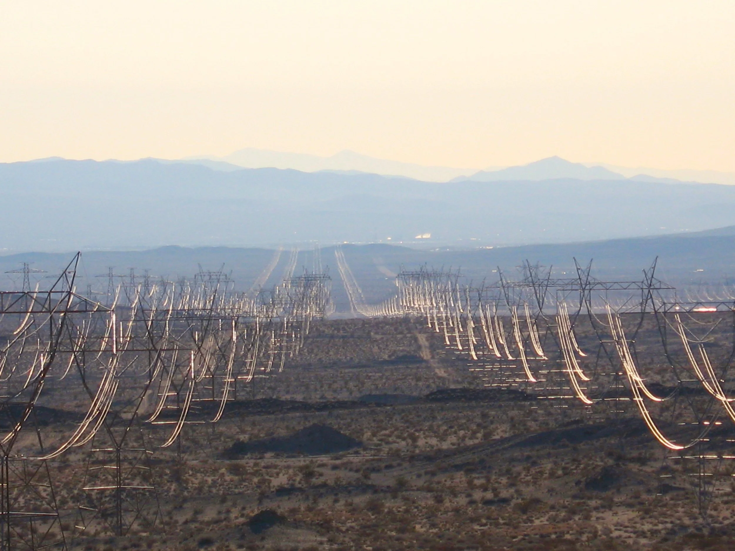 A landscape view of distant mountains, with power lines and electrical infrastructure in the foreground, taken during dawn or dusk with a hazy sky.