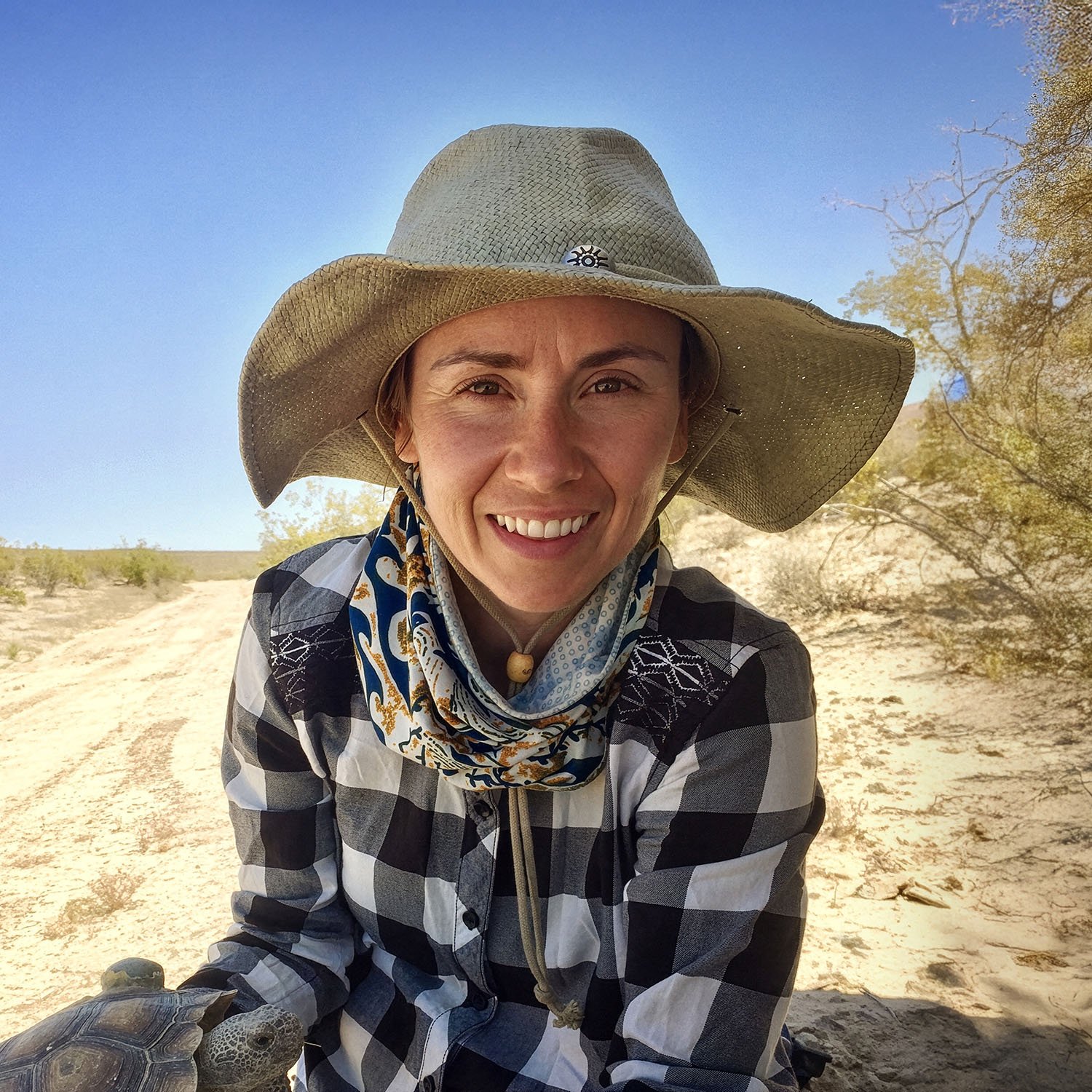 A woman wearing a wide-brimmed hat, a scarf, and a checkered shirt, smiling outdoors in a desert landscape with a tortoise in her hand.